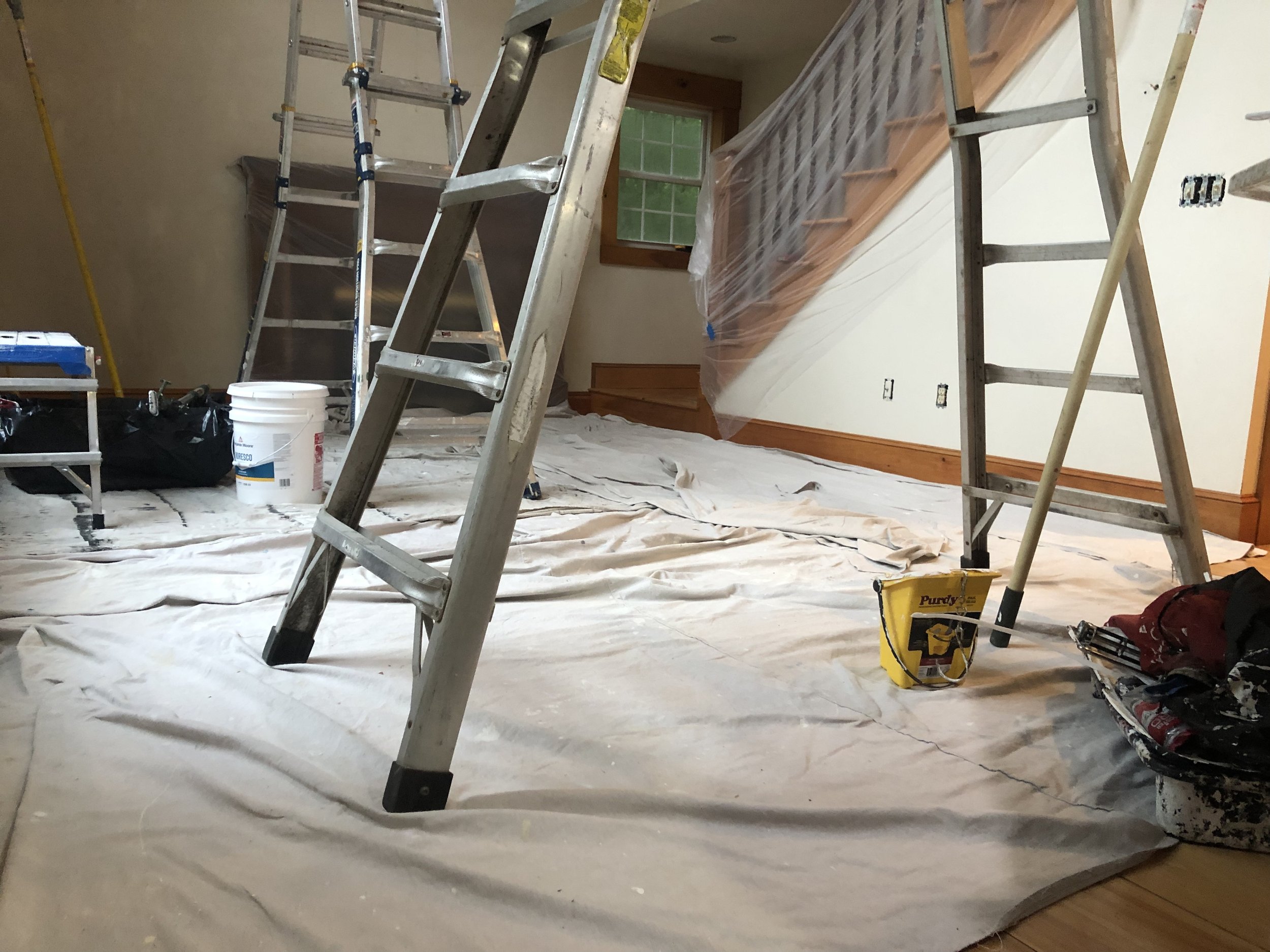Room under renovation with ladders, painter's tape, and painting supplies on the floor.