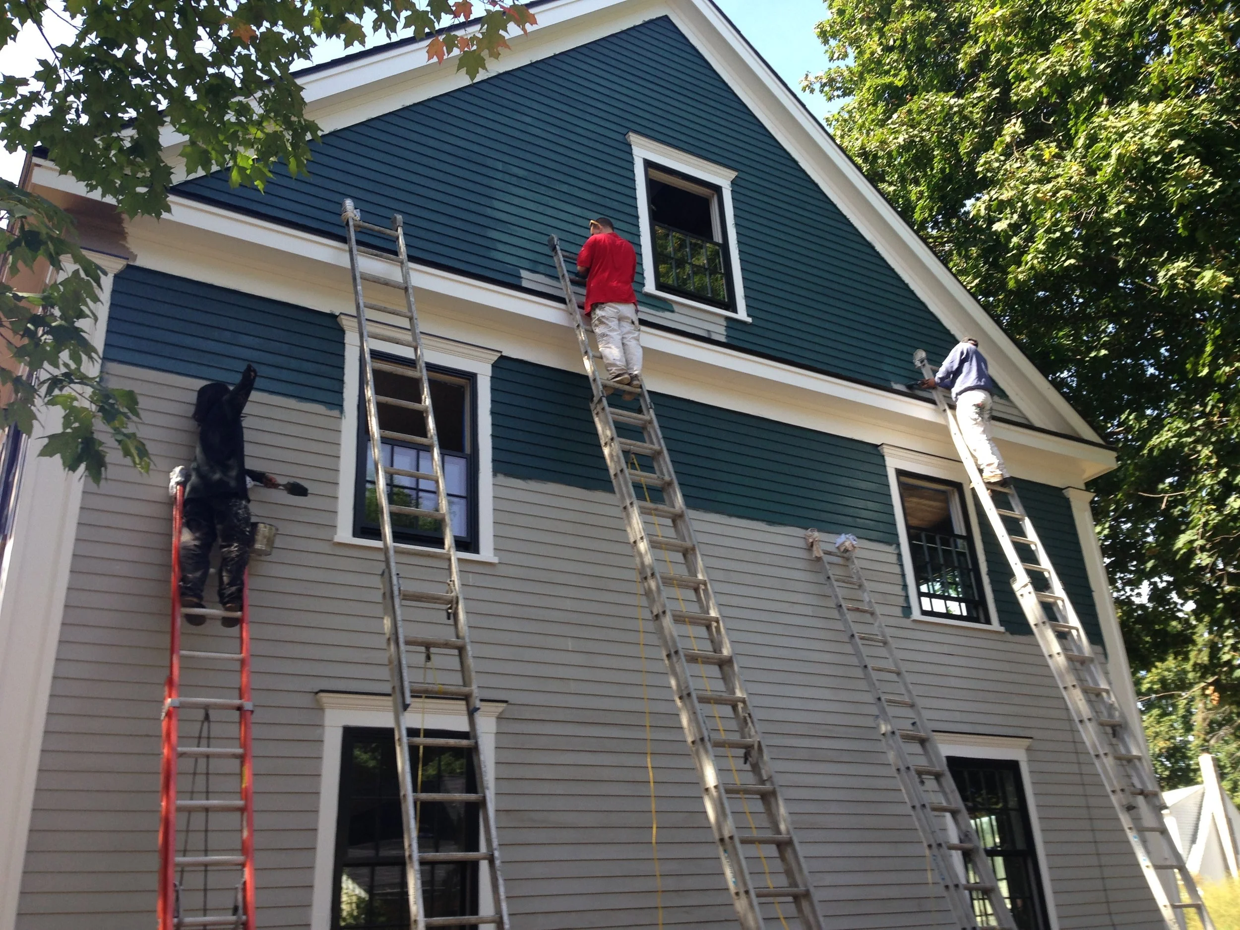 Three workers on ladders painting the exterior of a house with blue and white siding, surrounded by green trees.