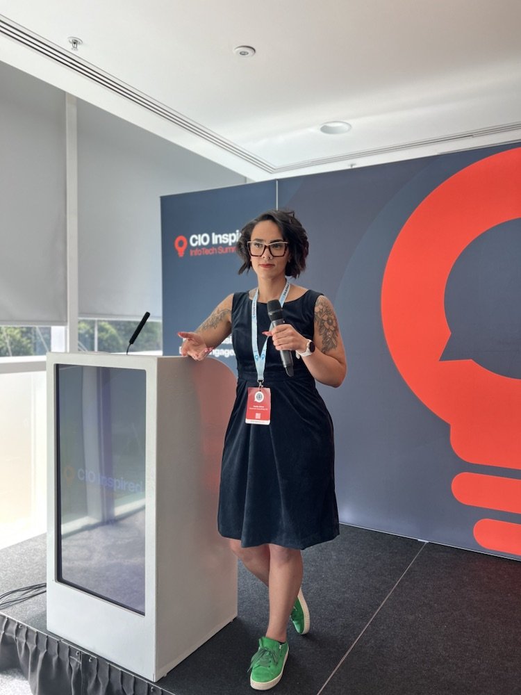 A woman with short dark hair, glasses, and tattoos on her arms, wearing a black dress and green sneakers, giving a presentation at a conference with a microphone. Behind her is a large logo with a red and black color scheme, and a screen displaying some text.
