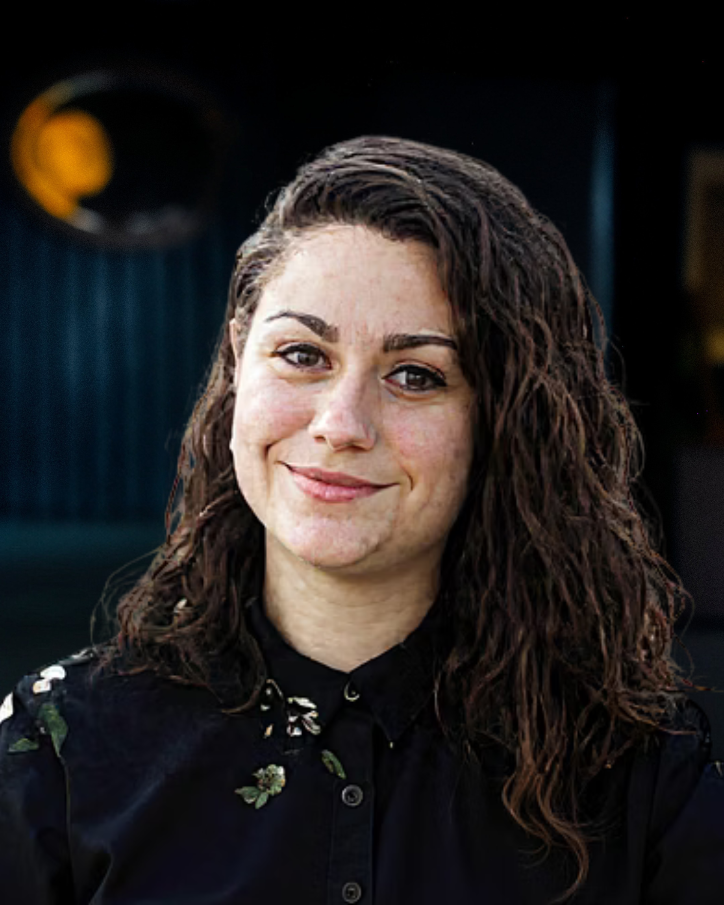 A woman with curly, dark brown hair and light skin smiling at the camera, wearing a black shirt with floral embroidery, standing indoors with dark blue walls and a yellow sign in the background.