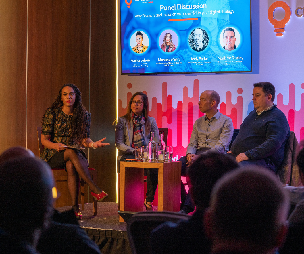 Panel discussion with four women and one man on stage at a conference, with a screen displaying speakers' names and titles behind them.