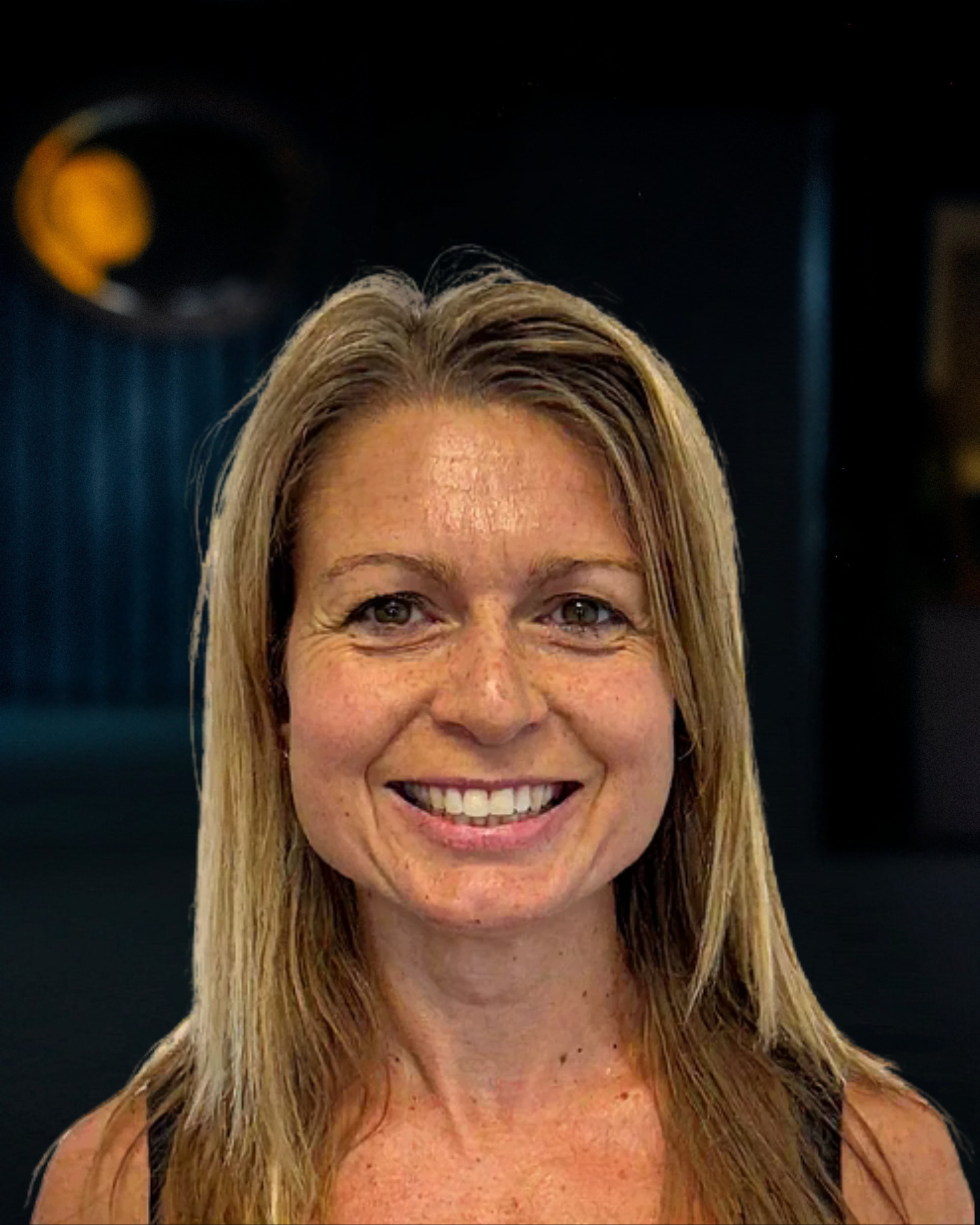 Close-up of a woman with blonde hair smiling at the camera, in an indoor setting.