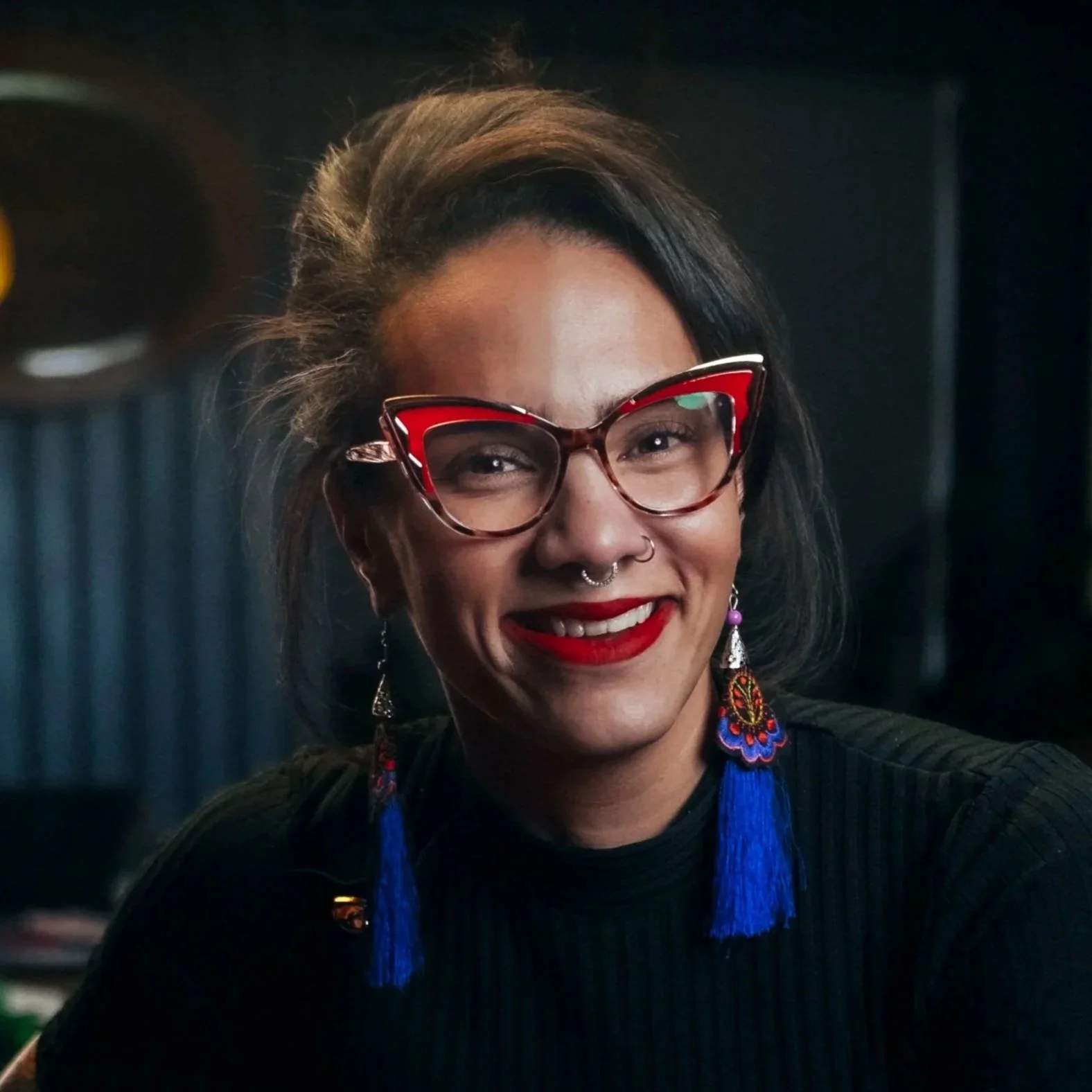 A woman with dark hair, wearing red and black cat-eye glasses, red lipstick, and colorful earrings with blue tassels, smiling at the camera.