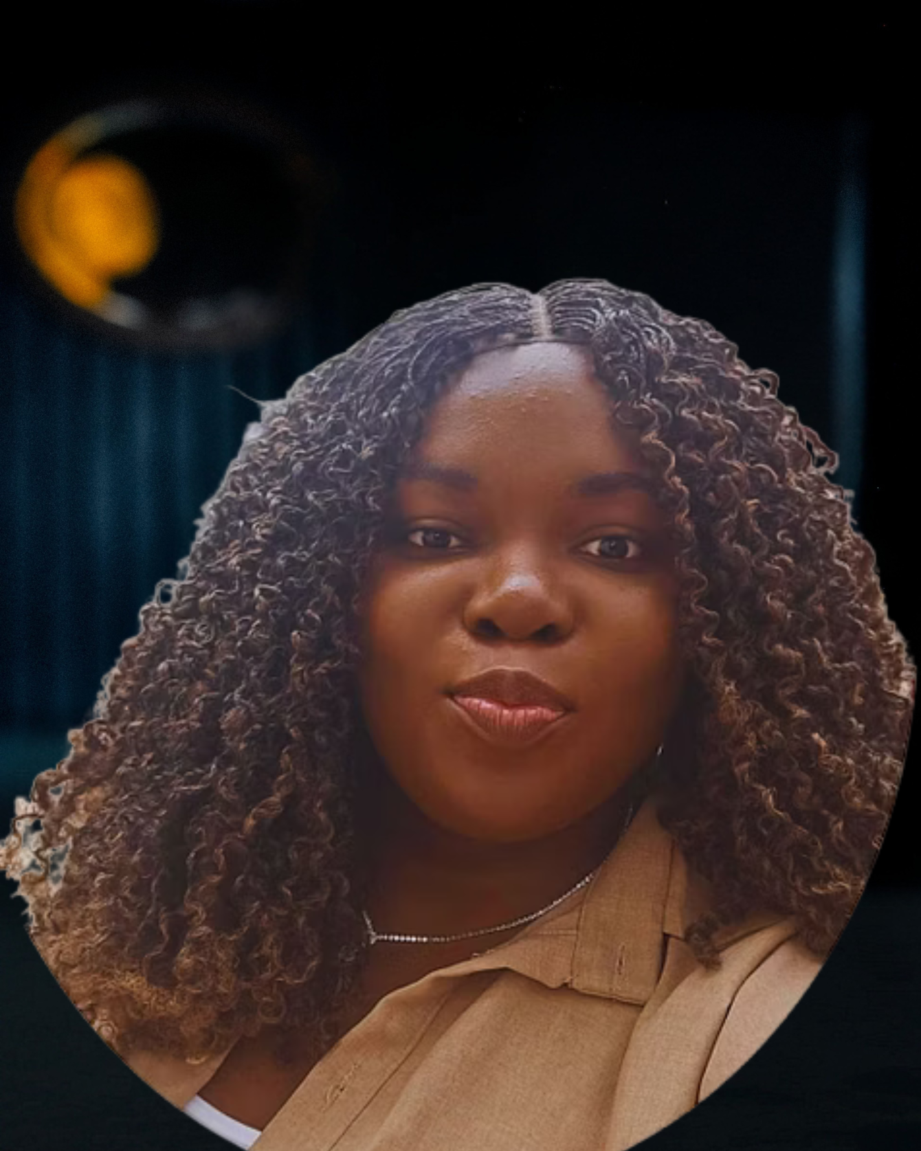 A woman with curly brown hair and medium skin tone posing indoors with a dark background and a blurred yellow and black circular object in the distance.