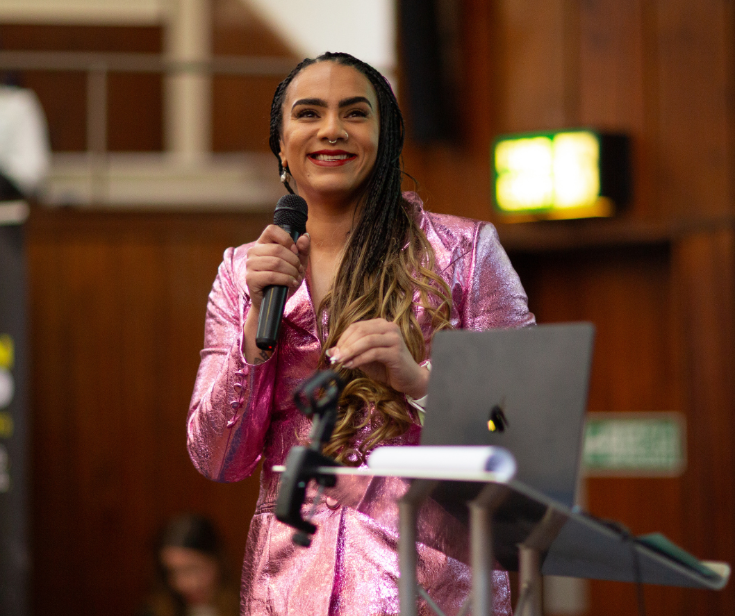 A woman in a shiny pink blazer smiling and holding a microphone at a presentation or event.