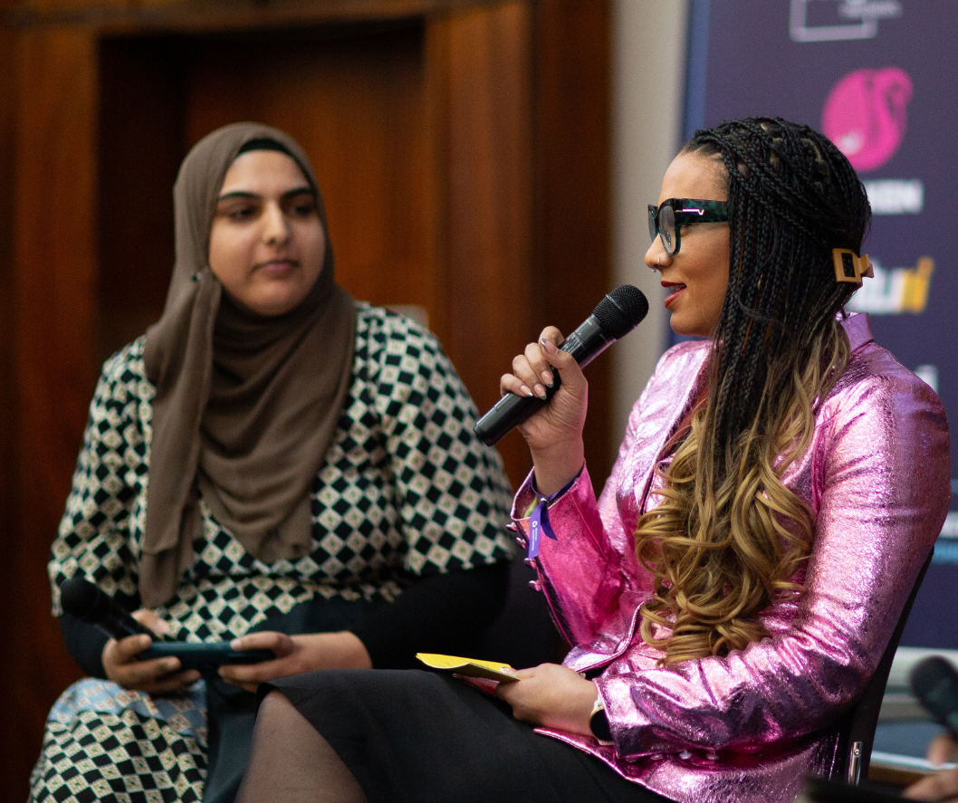 Two women are participating in a panel discussion or interview; one woman with long braided hair, wearing glasses and a pink shiny blazer, is speaking into a microphone. The other woman, wearing a checkered dress with a hijab, is holding a tablet and listening.