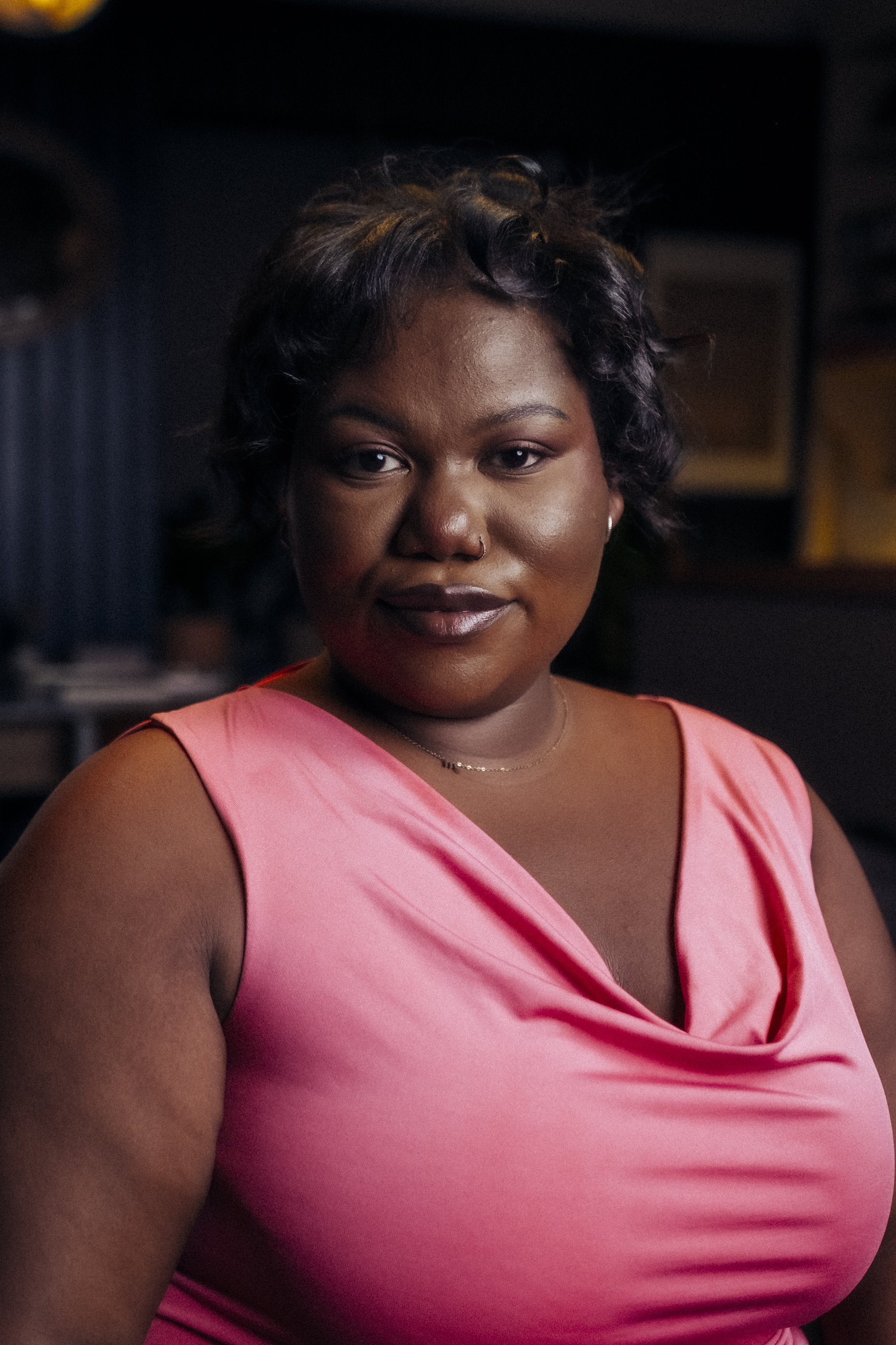 A woman with dark hair styled in curls, wearing a pink sleeveless top, smiling softly, with a blurred background.