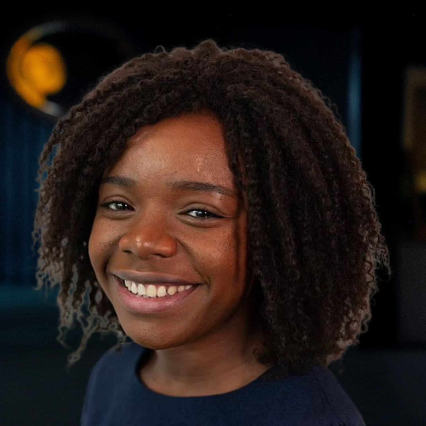 Close-up of a smiling young woman with curly dark hair, wearing a dark top, in an indoor setting.
