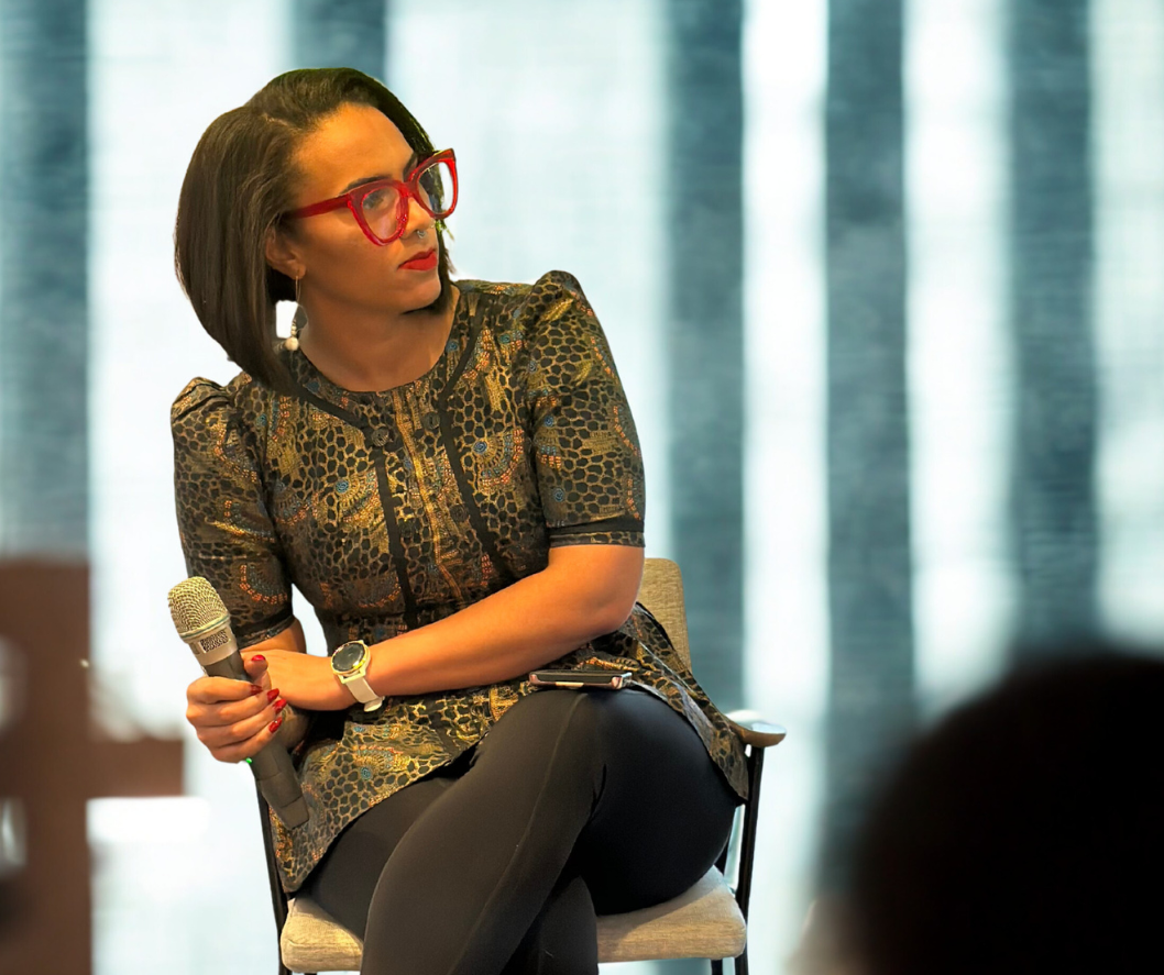 A woman with short dark hair, wearing red glasses and a patterned top, sitting on a chair holding a microphone, with a serious expression in front of a curtain.