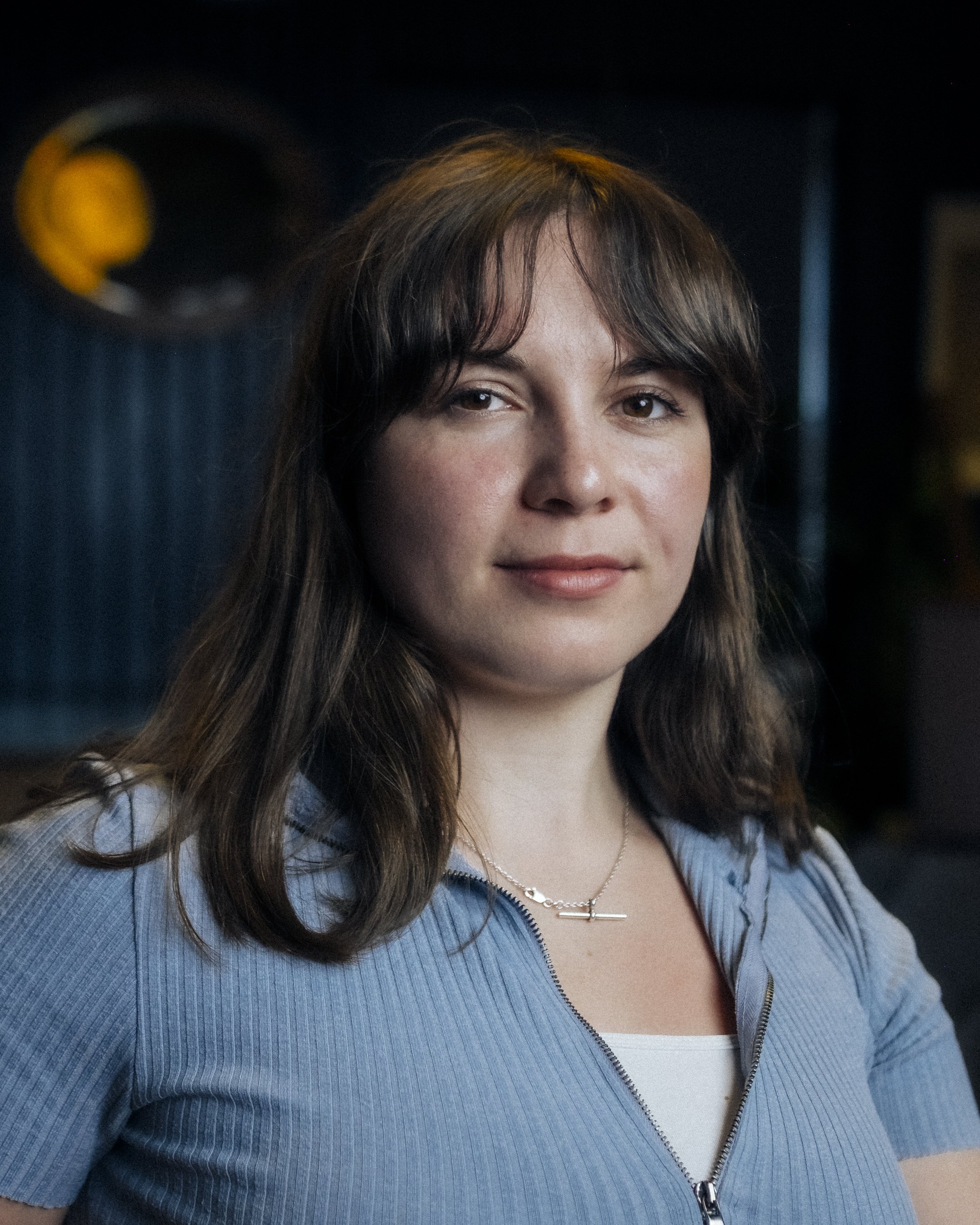 A woman with shoulder-length brown hair and bangs, wearing a light gray zip-up top and a silver chain necklace, smiling softly indoors.