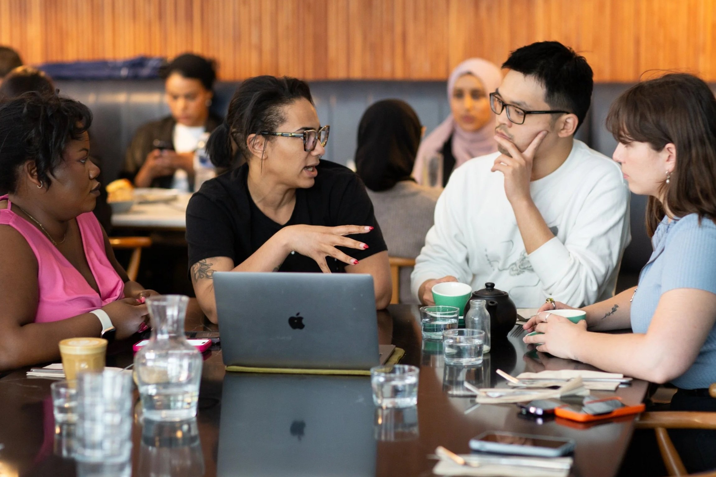 A diverse group of women and a man engaged in a discussion around a conference table with drinks, notebooks, and a laptop.