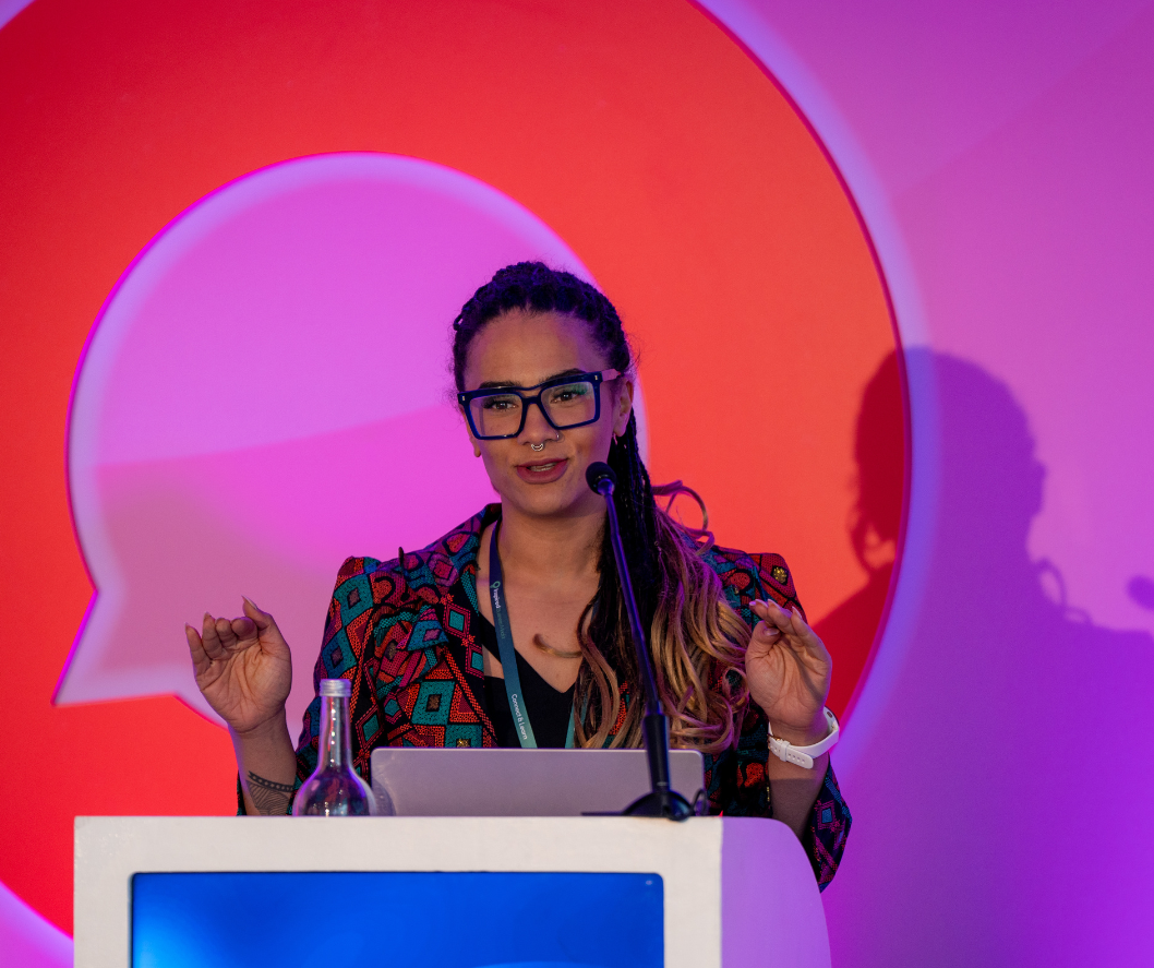 A woman with glasses and styled hair is speaking at a conference behind a white podium. There is a laptop, a water bottle, and a microphone on the podium. The background features a large speech bubble graphic with purple and red lighting.
