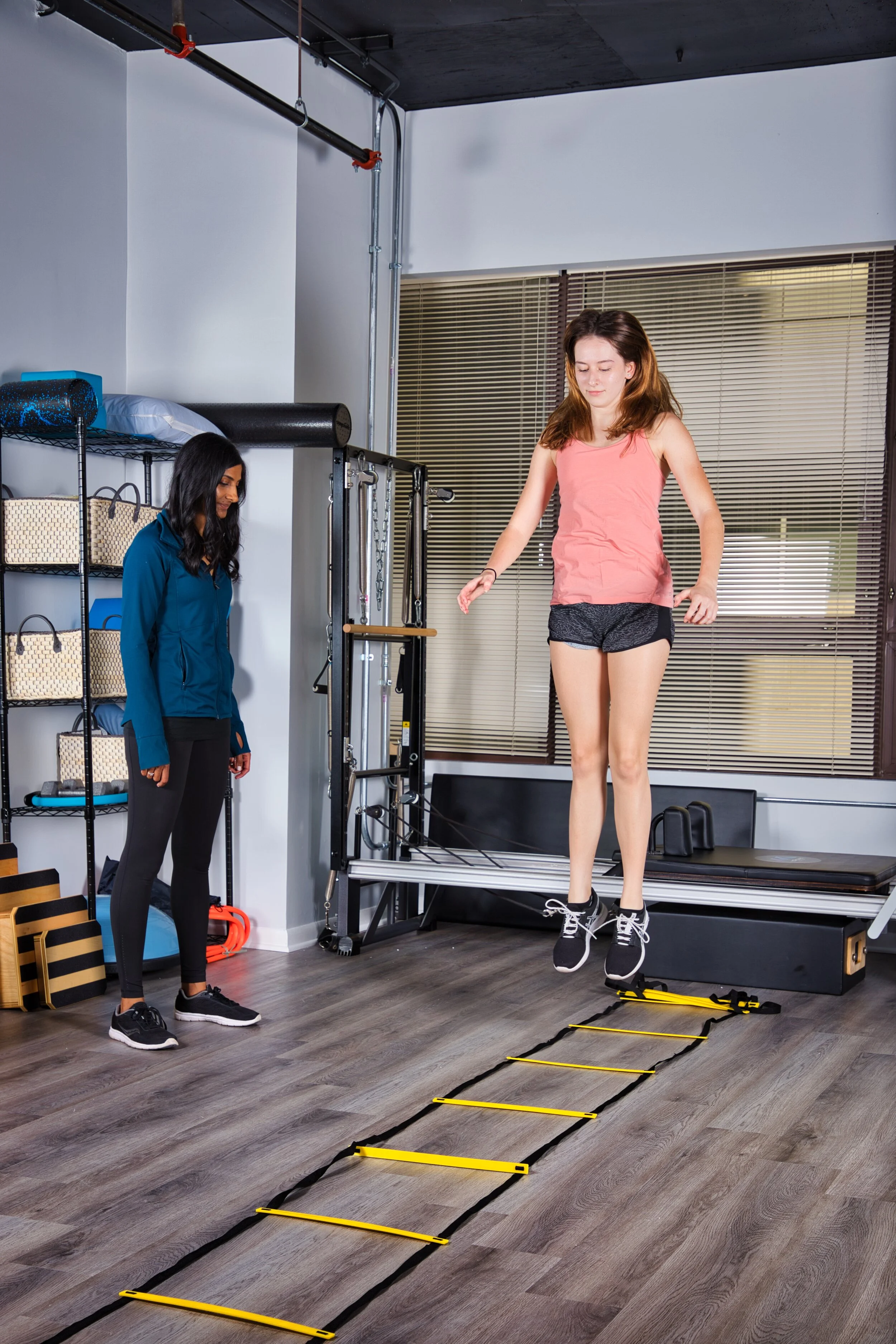 A young girl practicing agility drills on a yellow training ladder in a gym, supervised by an physical therapist.
