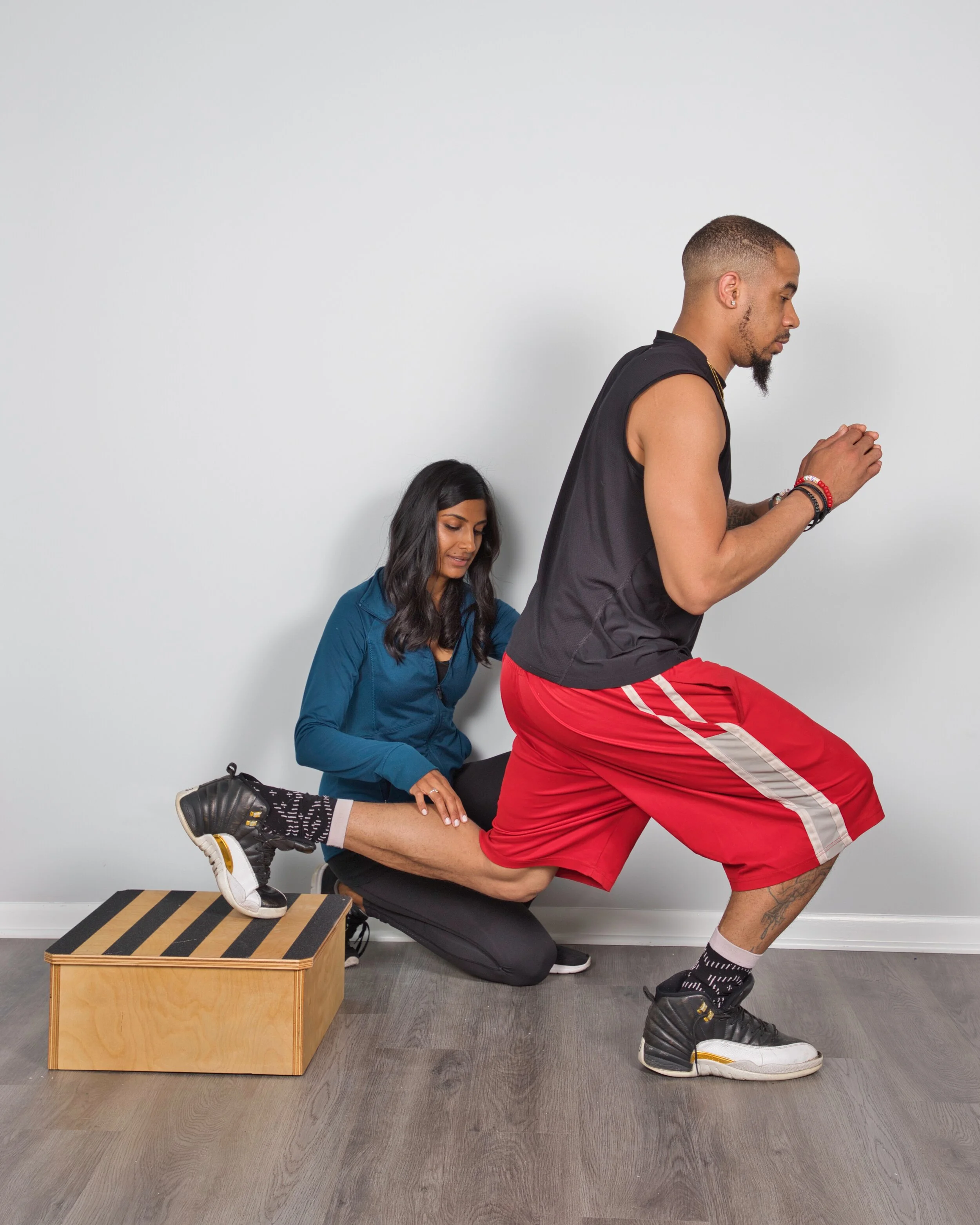 A man doing a squat exercise with a woman helping him, holding his ankle, on a wooden box. They are in a gym or fitness space with a plain white wall.