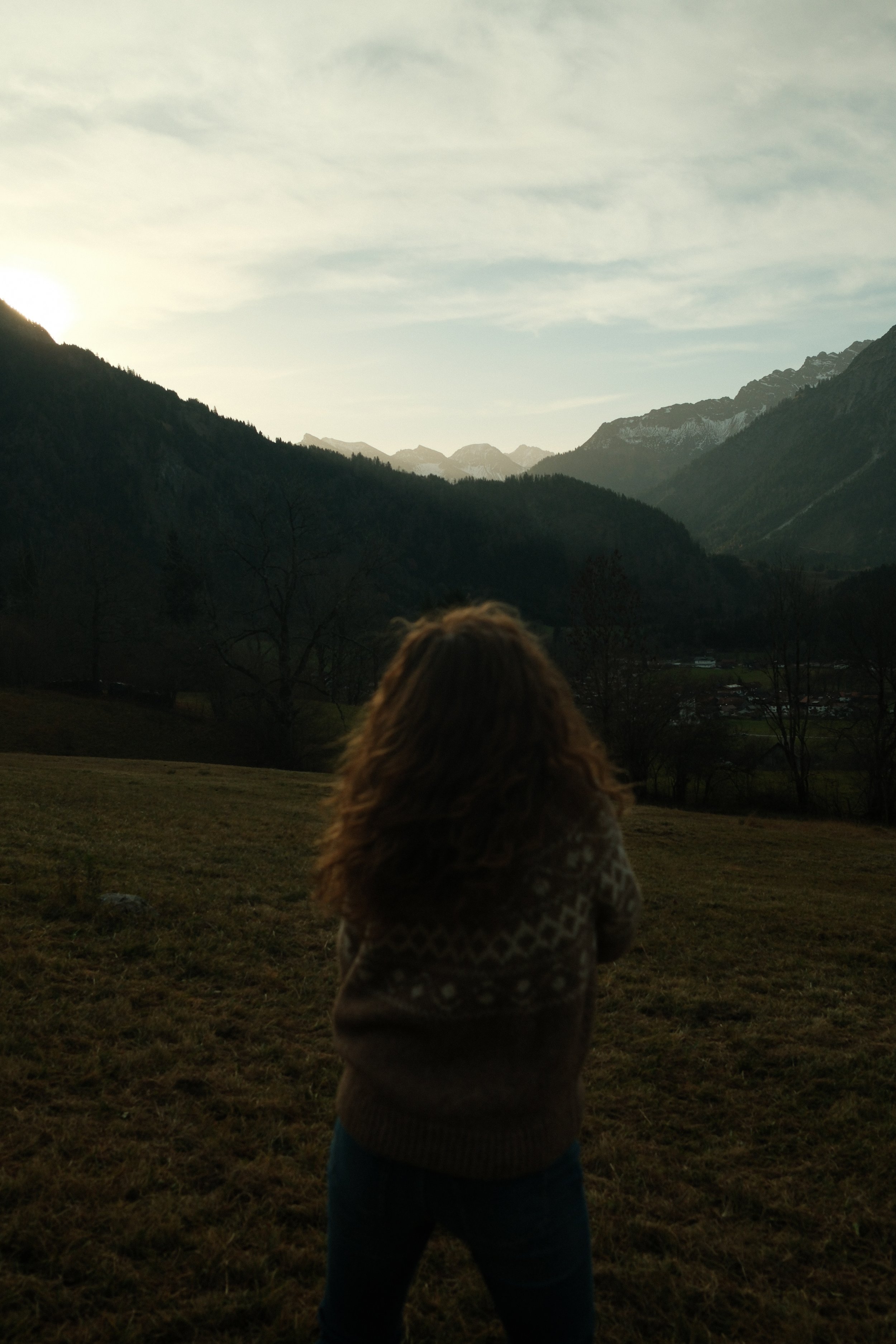 Ein Mädchen mit lockigem Haar steht auf einer Wiese und blickt auf eine Berglandschaft bei Sonnenuntergang.