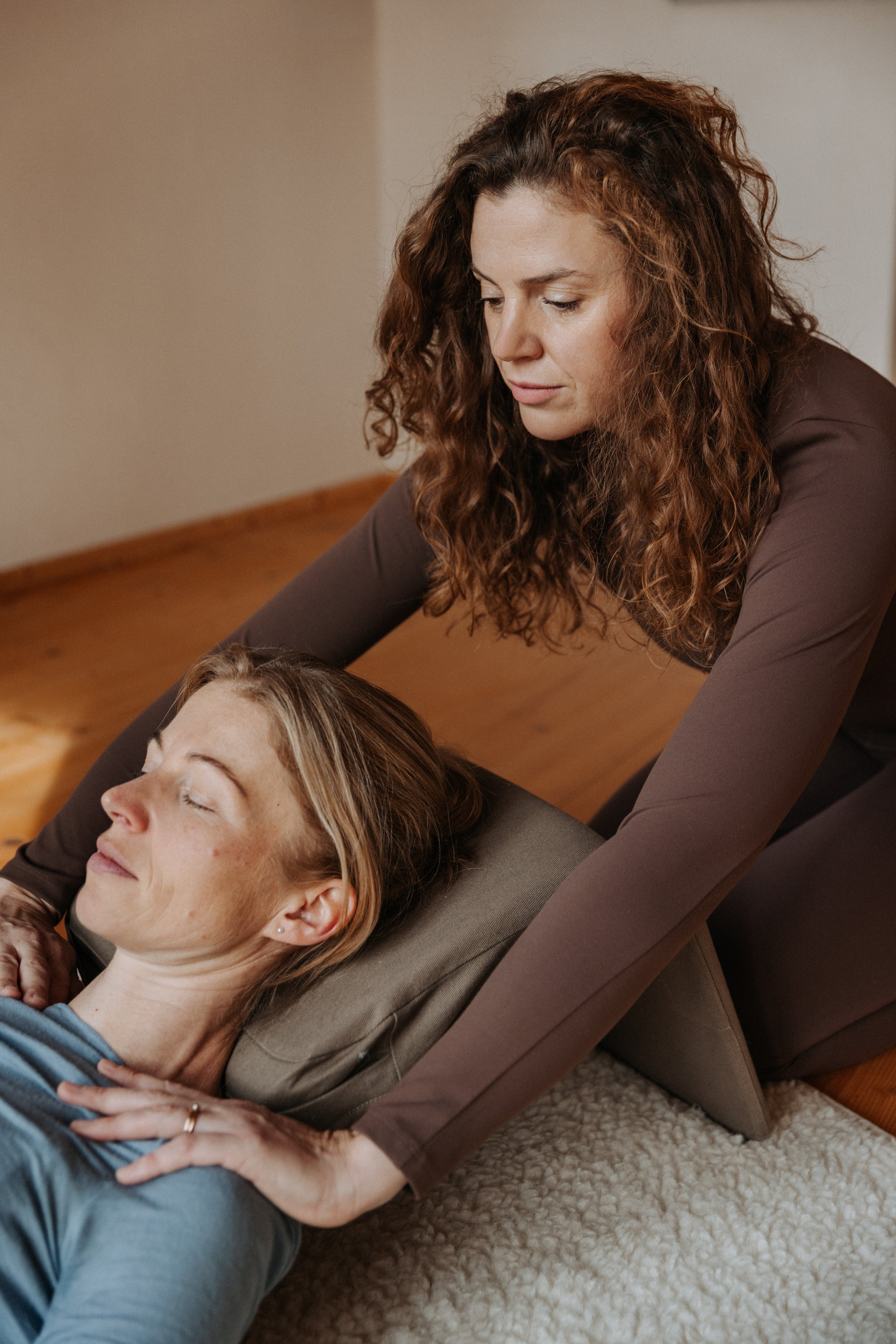 Zwei Frauen, eine liegend auf einem Rücken mit geschlossenen Augen, die andere darüber gebeugt, offenbar in einer entspannenden oder therapeutischen Situation in einem ruhigen Raum.