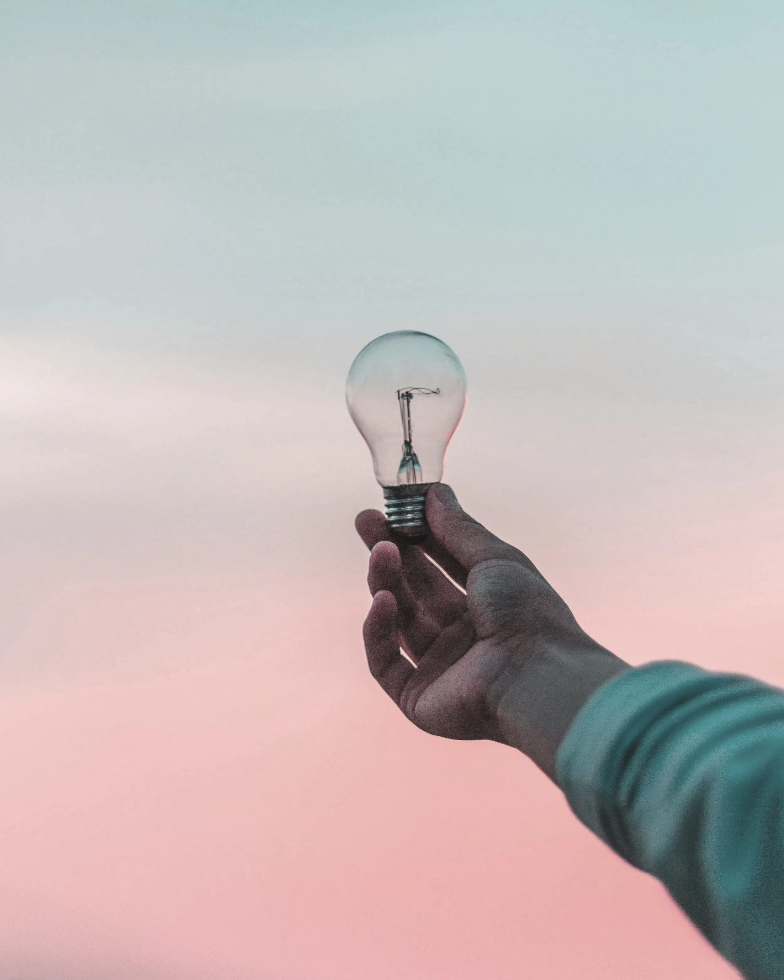 Hand hält eine Glühbirne vor einem Pastellfarbenen Himmel.