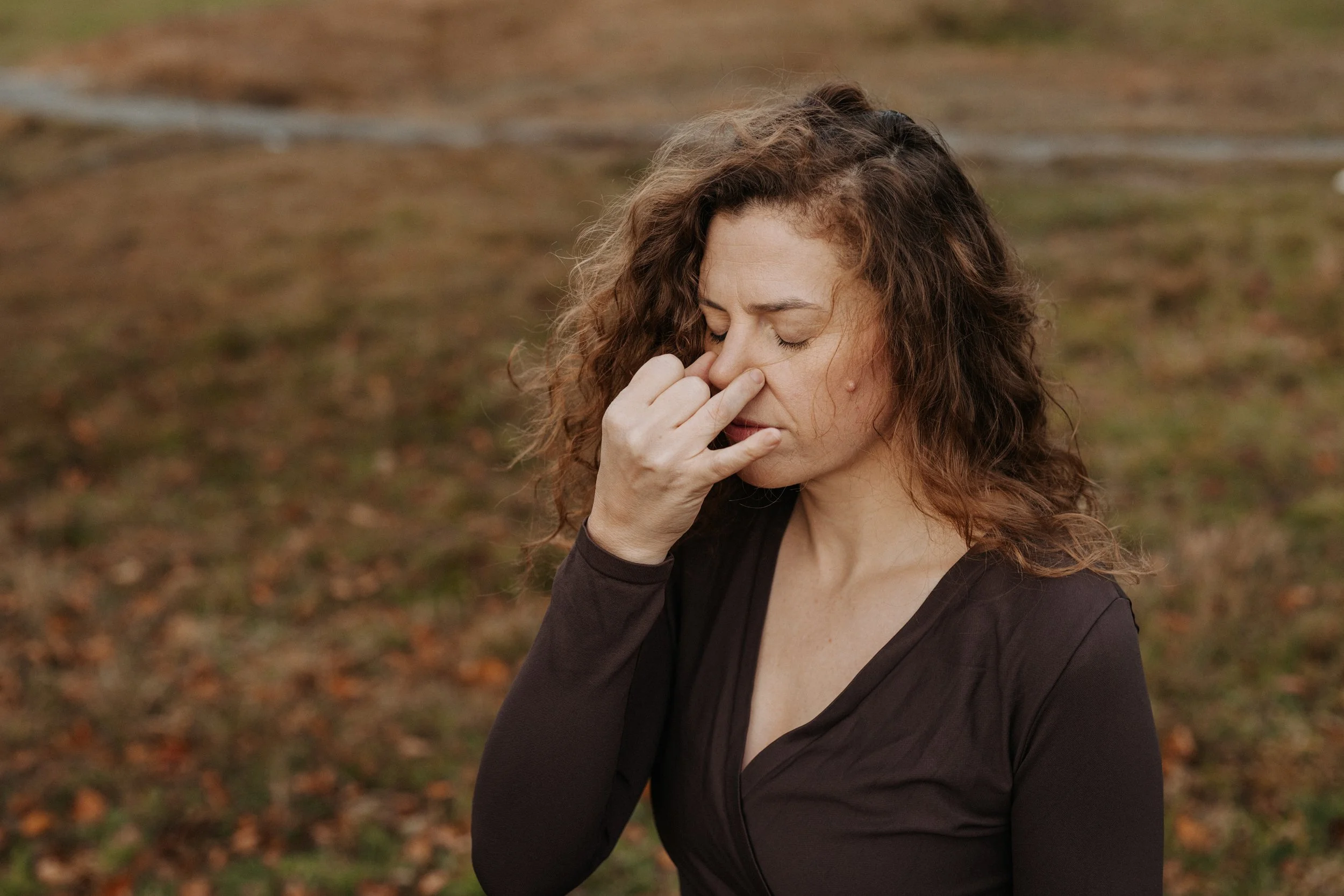 Eine Frau mit lockigem Haar steht draußen in einem herbstlichen, blätterbedeckten Bereich, und praktiziert die Wechselatmung.