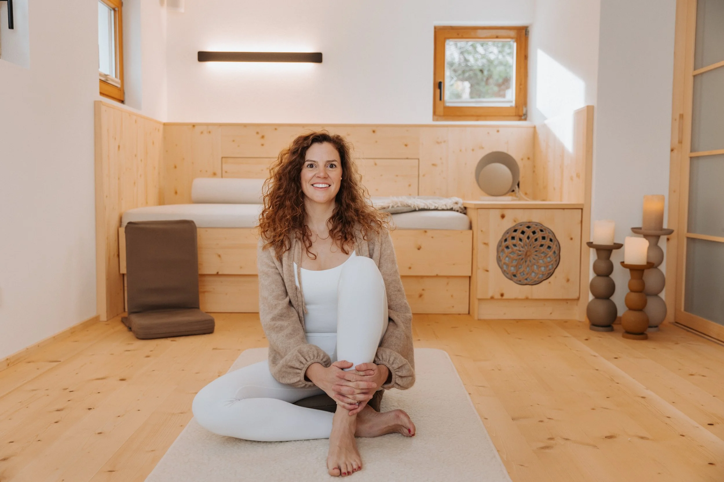 Eine lachende Frau sitzt auf einem hellen Teppich vor einem Bett in einem Raum mit Holzverkleidung und großer Fensterwand. Sie hält ein großes, weißes, ovales Kissen im Schoß. Sie befindet sich in ihren eigenen Praxisräumen.