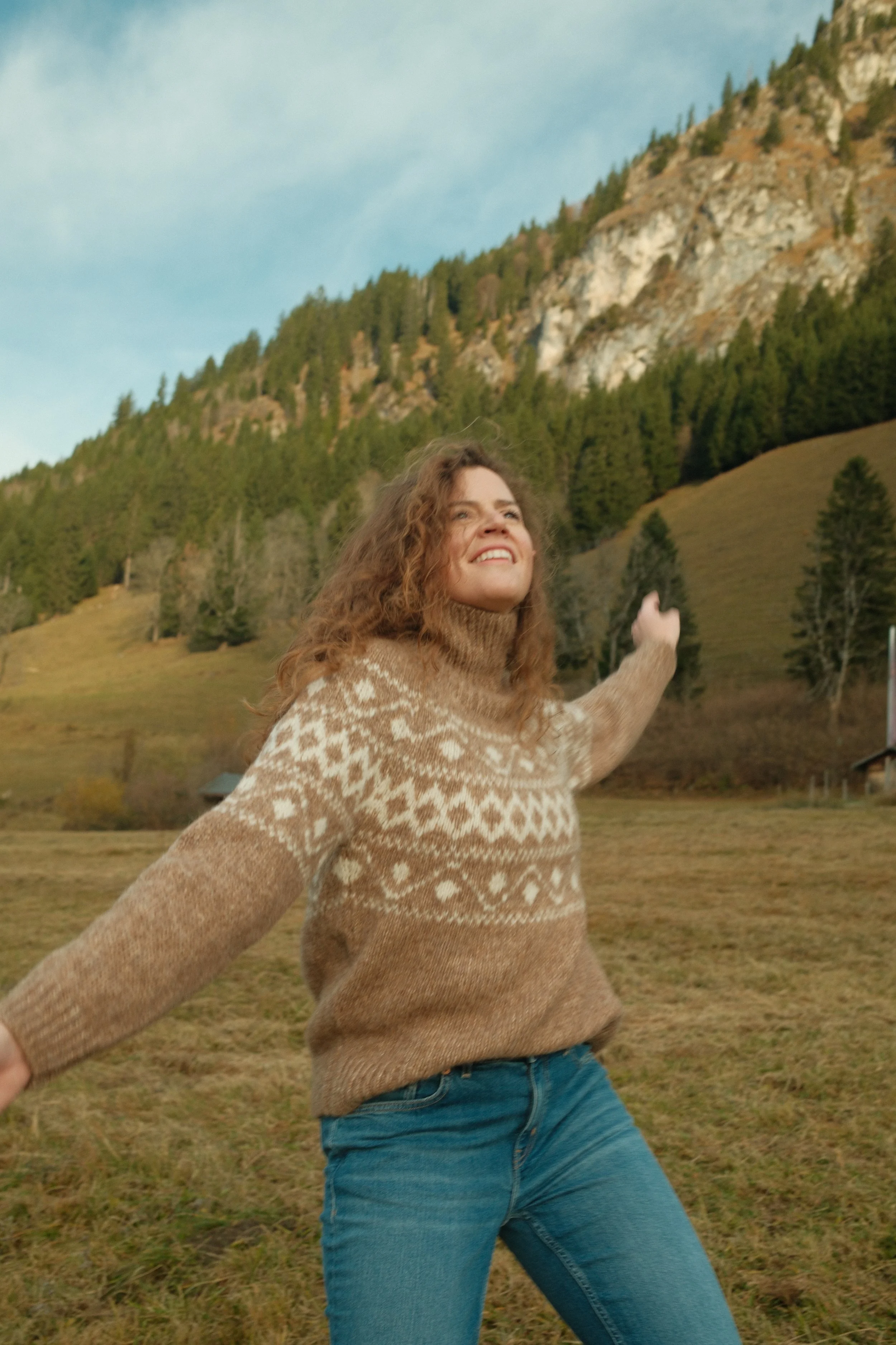 Frau in buntem Pullover und Jeans, die in einer offenen Landschaft mit Hügeln und Bäumen steht, lächelnd und die Arme ausgebreitet.