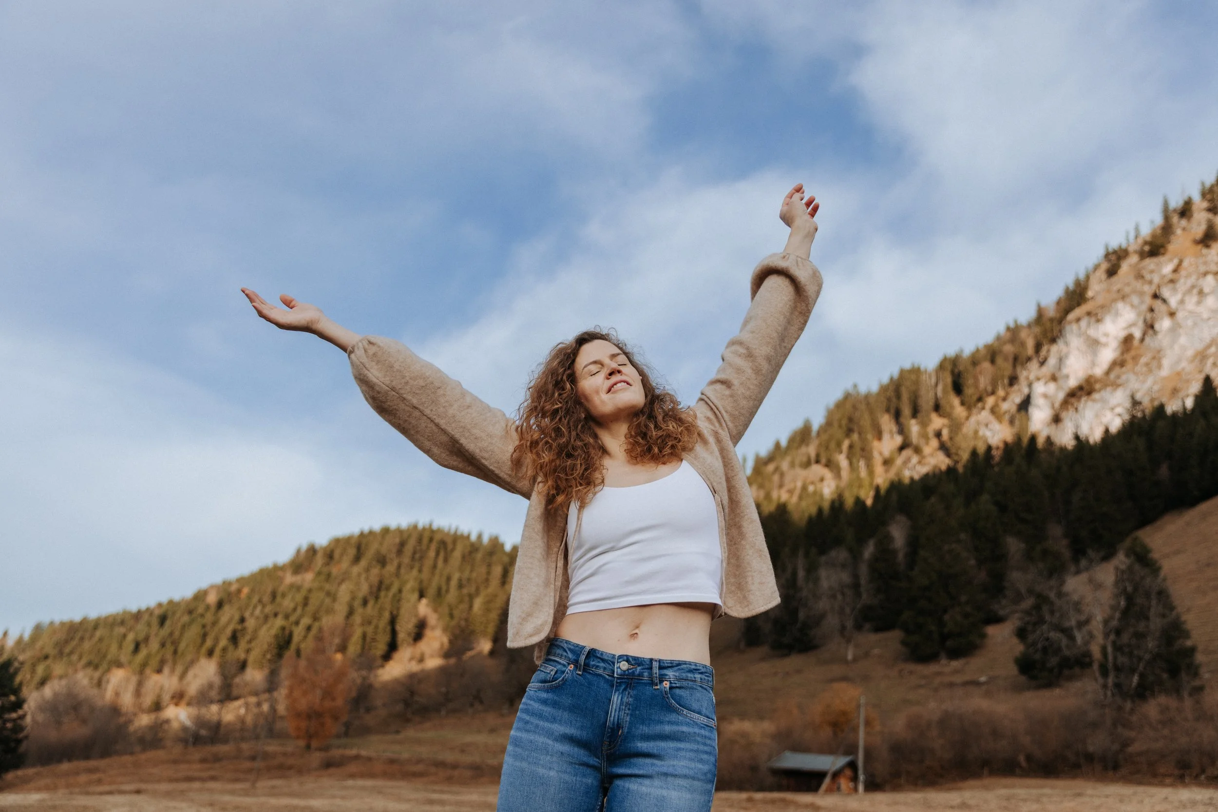 Junge Frau mit offenen Armen steht in der Natur, umgeben von Hügeln und Bäumen, unter einem blauen Himmel.