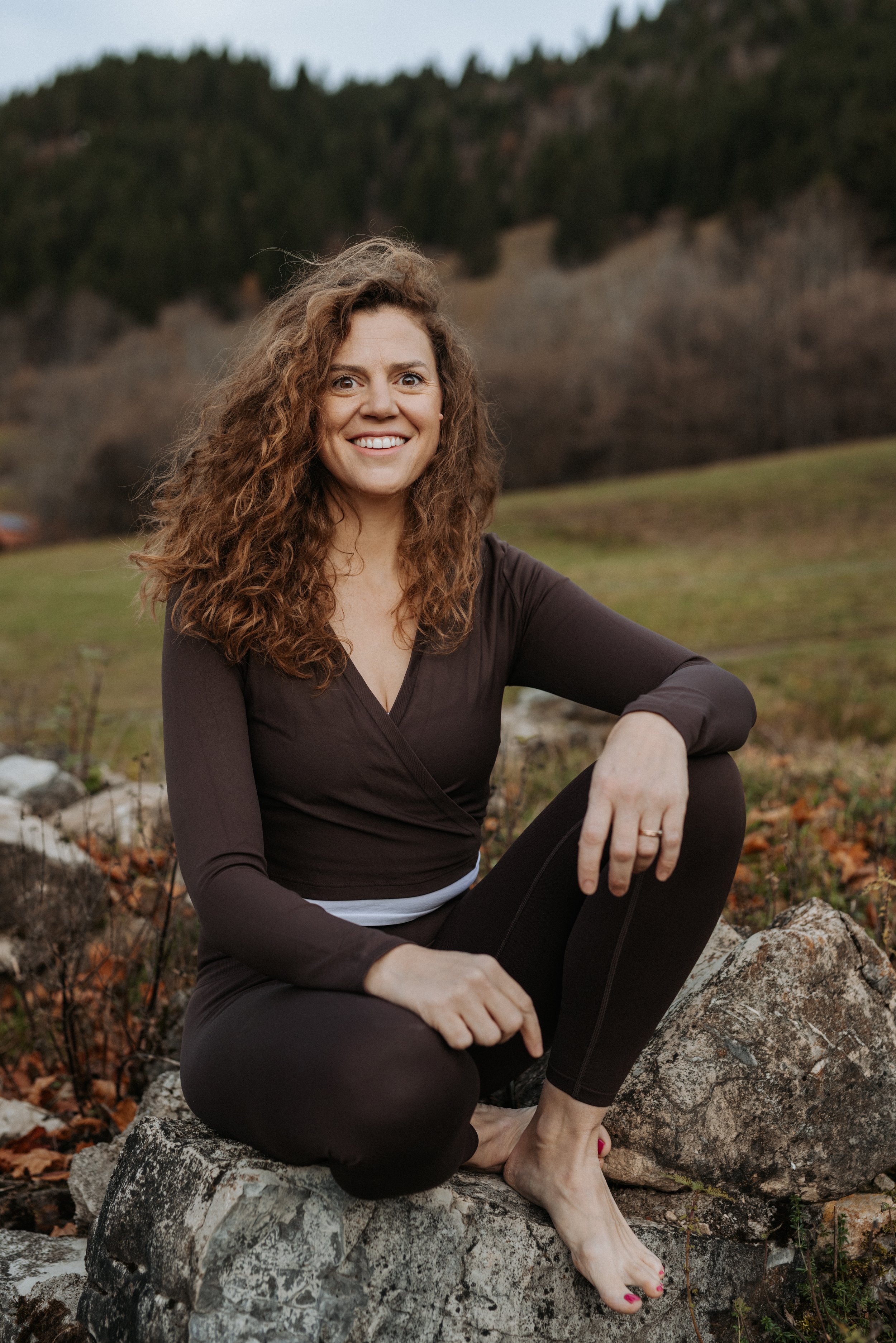 Eine lächelnde Frau mit lockigem braunem Haar sitzt barfuß auf einem Felsen im Freien, umgeben von einer natürlichen Landschaft mit Hügeln und Bäumen im Hintergrund.