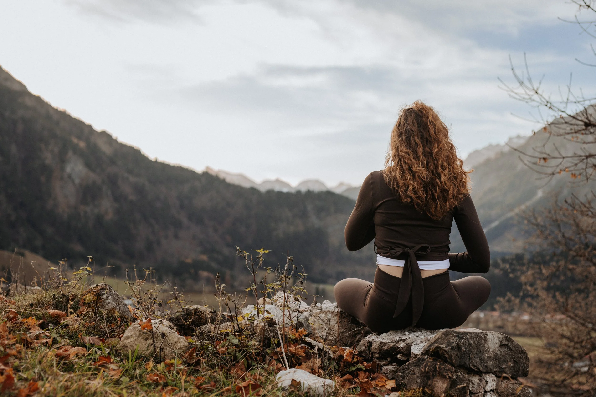 Frau sitzt während einer Atemübung auf einem Felsen und blickt auf eine Berglandschaft, trägt schwarze legging und Oberteil, mit lockigem braunem Haar.