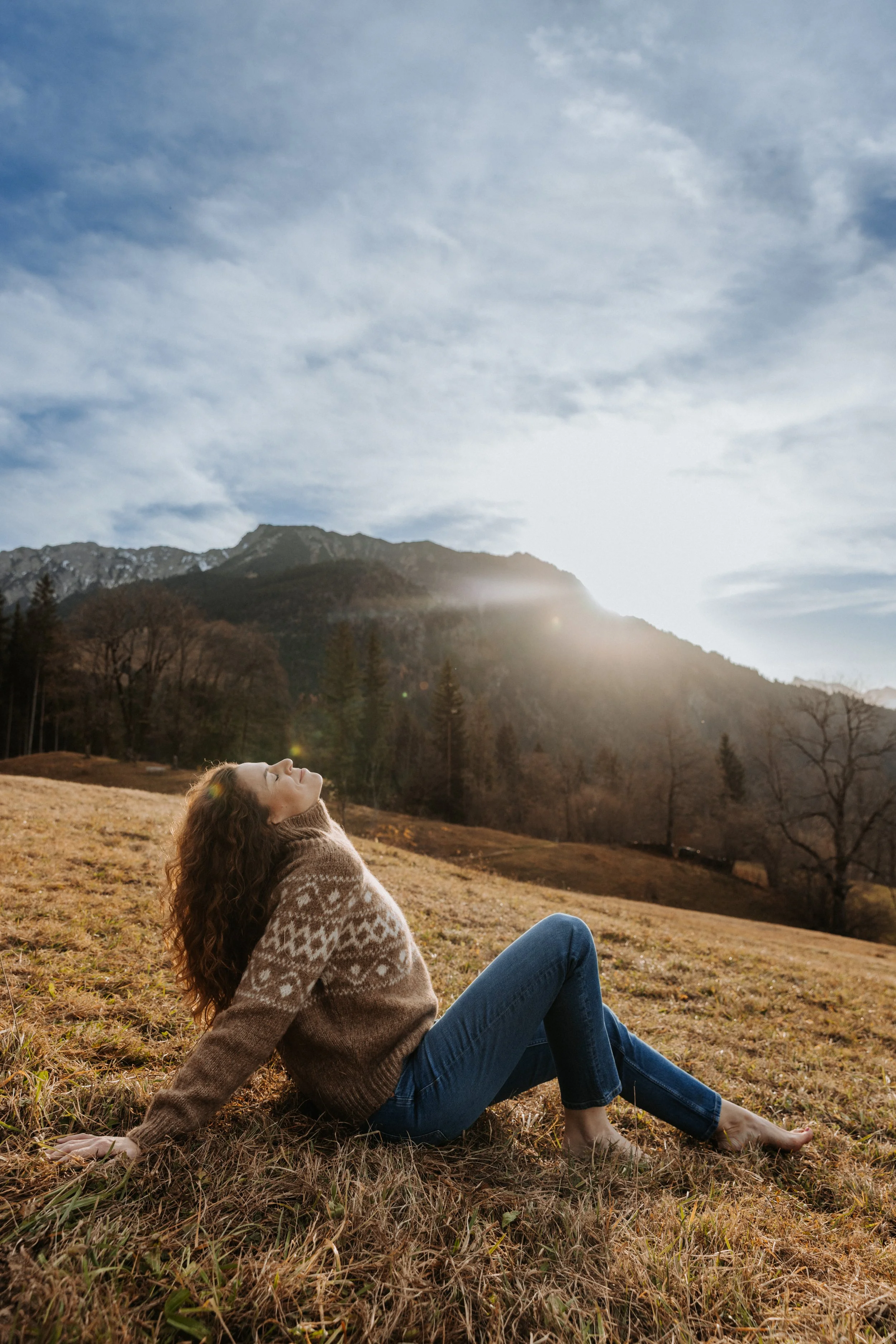 Frau sitzt entspannt auf einer Wiese mit Bergen im Hintergrund, Sonnenlicht und Himmel mit Wolken.