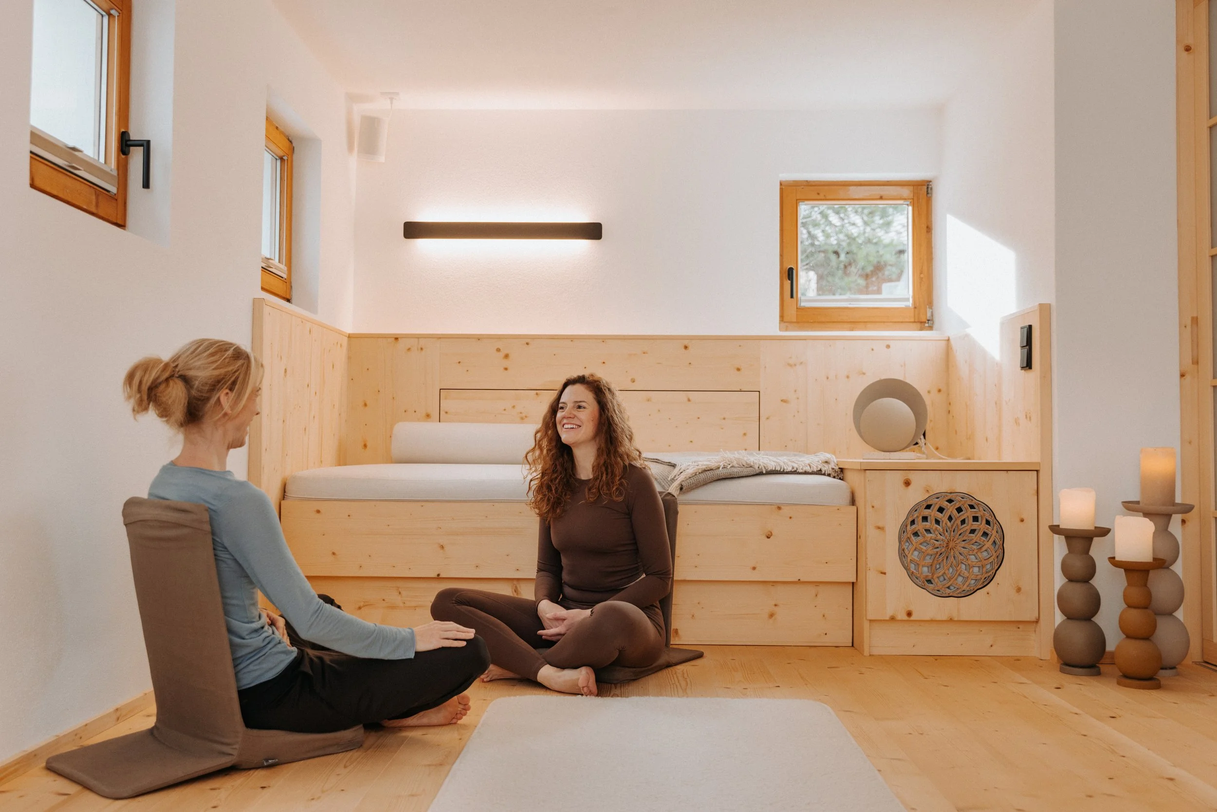 Zwei Frauen sitzen im Yoga-Raum, eine lächelt, die andere sitzt im Schneidersitz auf dem Boden, umgeben von Kerzen und Dekorationen.