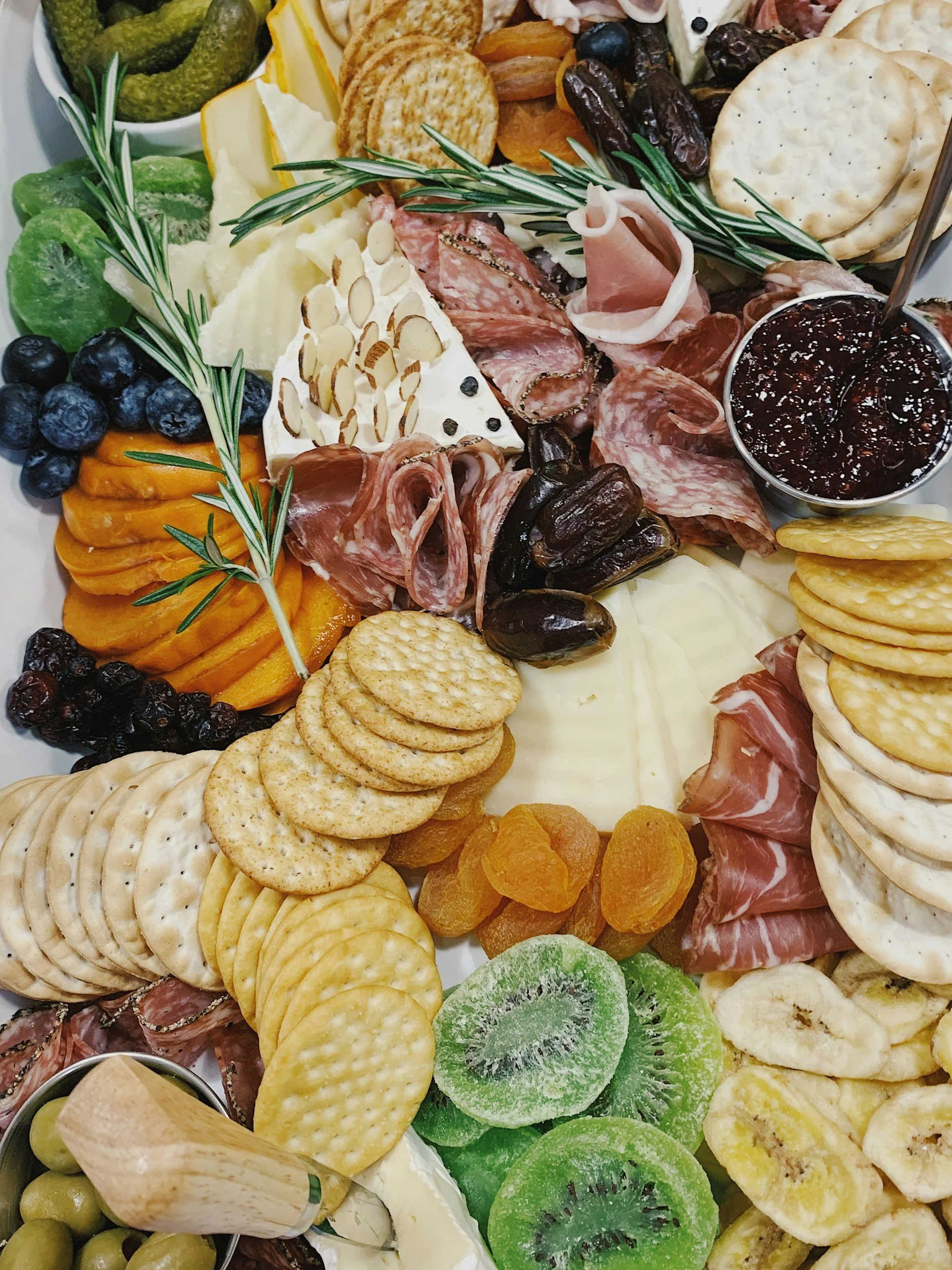 Charcuterie board with assorted cheeses, crackers, fresh and dried fruits, nuts, jam, and cured meats including prosciutto and salami, garnished with sprigs of rosemary from The Charcuterie Chics in Huntersville NC