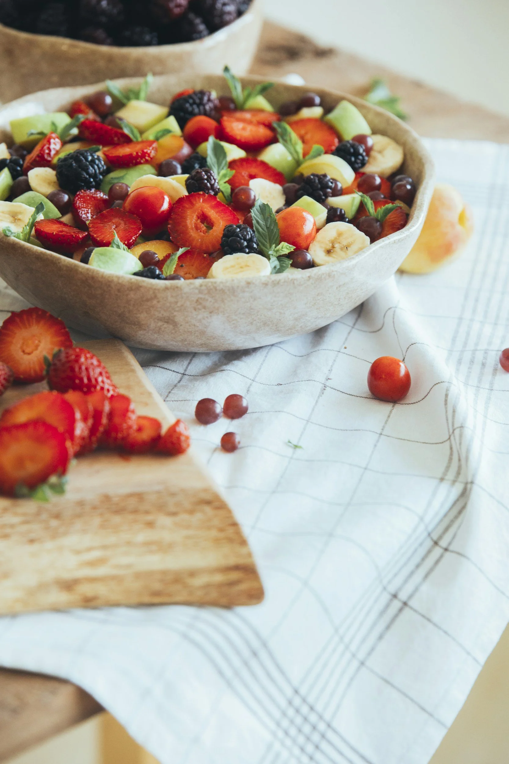 A bowl of mixed fresh fruit salad containing strawberries, blackberries, blueberries, grapes, banana slices, and mint leaves on a white checkered tablecloth with a cutting board of sliced strawberries nearby from The Charcuterie Chics near Asheville