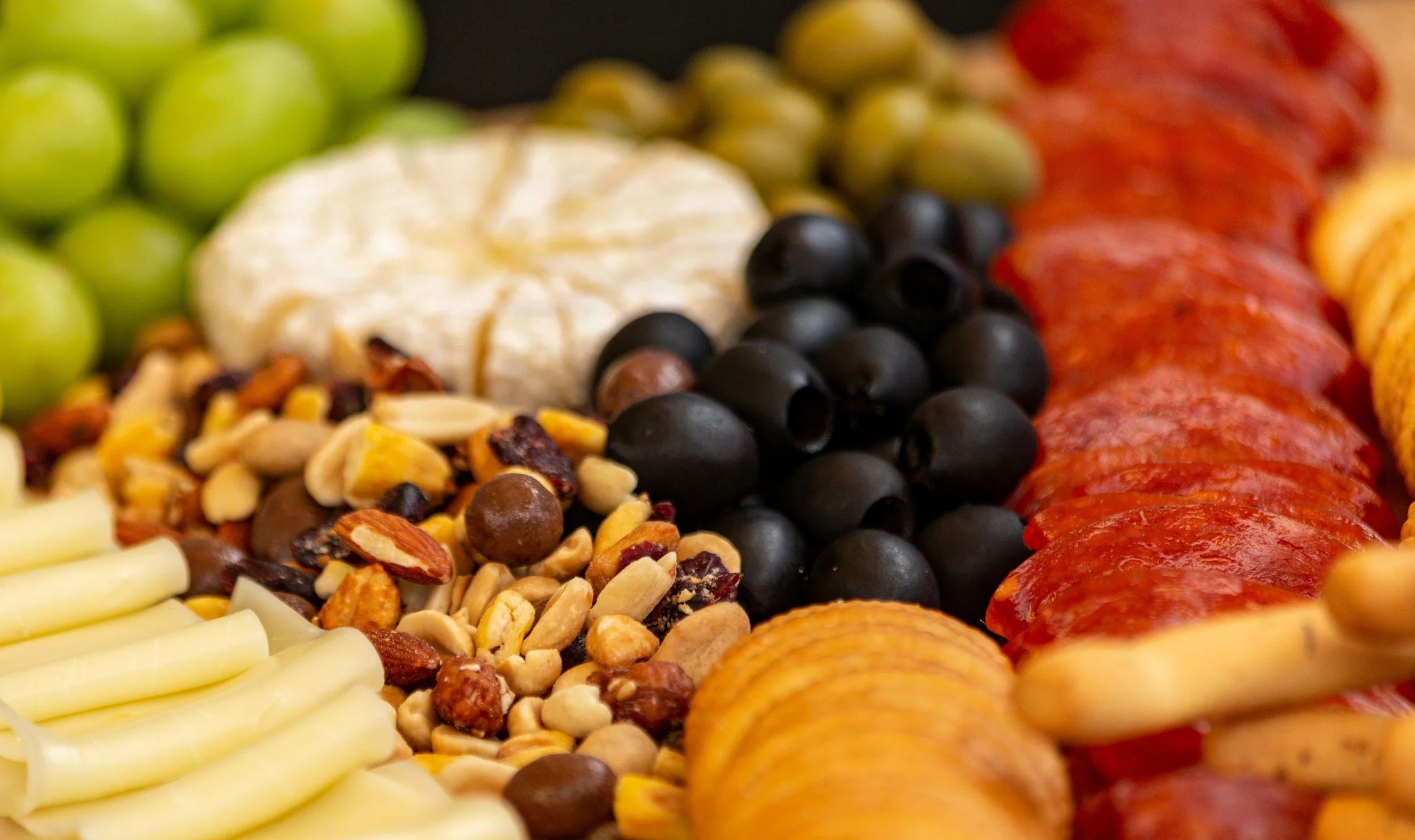 Close-up of assorted snacks including green grapes, a wheel of brie cheese, mixed nuts, black olives, salami slices, potato chips, and breadsticks from The Charcuterie Chics near Winston Salem NC