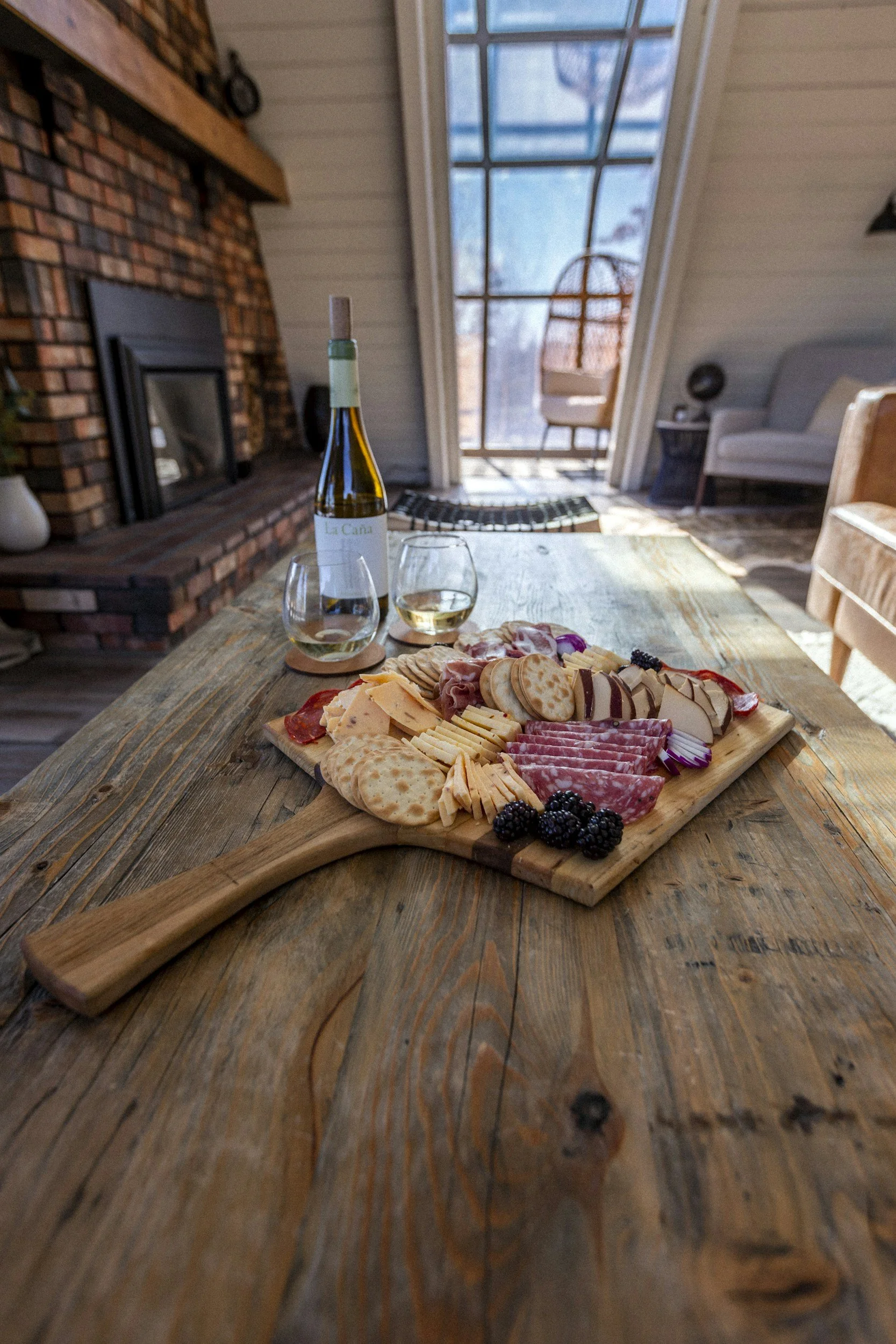 A cheese and charcuterie board with crackers and blackberries on a wooden table, with a bottle of white wine and two glasses in a cozy living room with a fireplace and a large glass door leading outside from The Charcuterie Chics near Huntersville NC