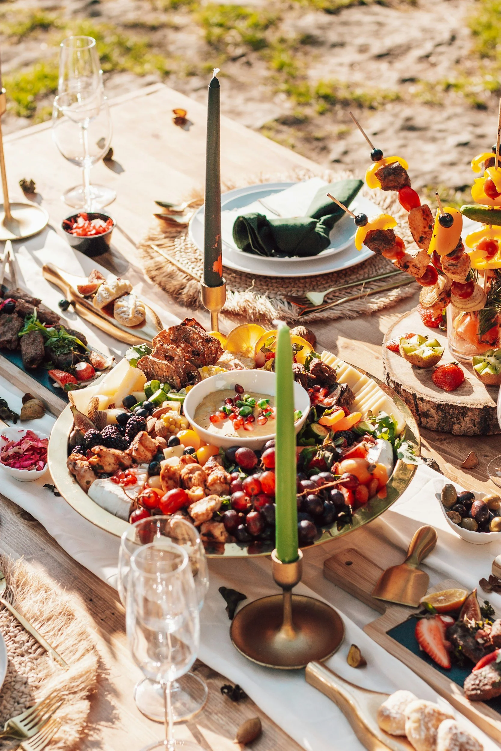 A festive outdoor dinner table decorated with colorful charcuterie foods, candles, wine glasses, and candles, set on a wooden table in natural light from The Charcuterie Chics near Raleigh NC
