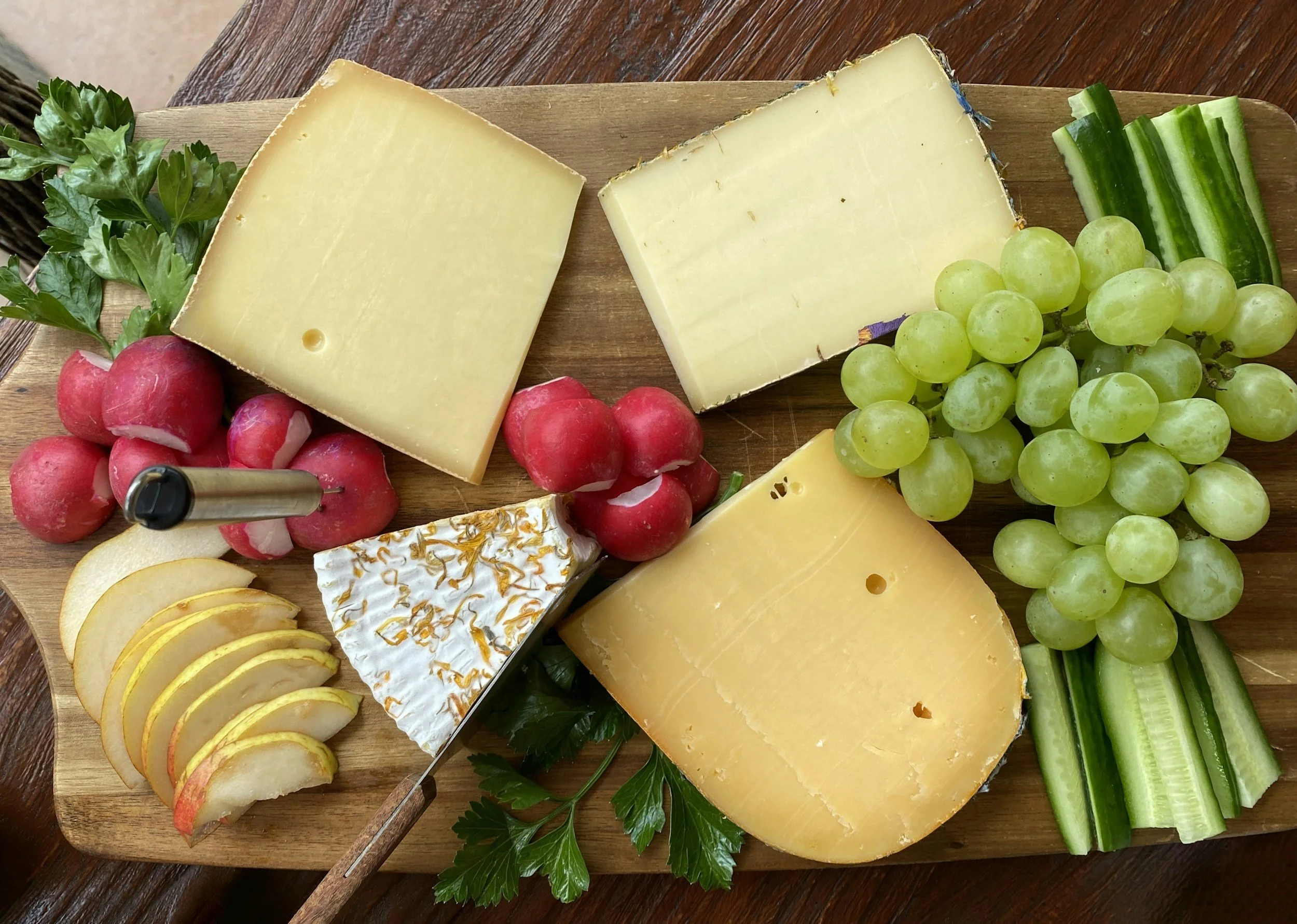A cheese platter with various cheeses, grapes, radishes, cucumber slices, sliced apple, and fresh parsley on a wooden board from The Charcuterie Chics in Winston Salem NC