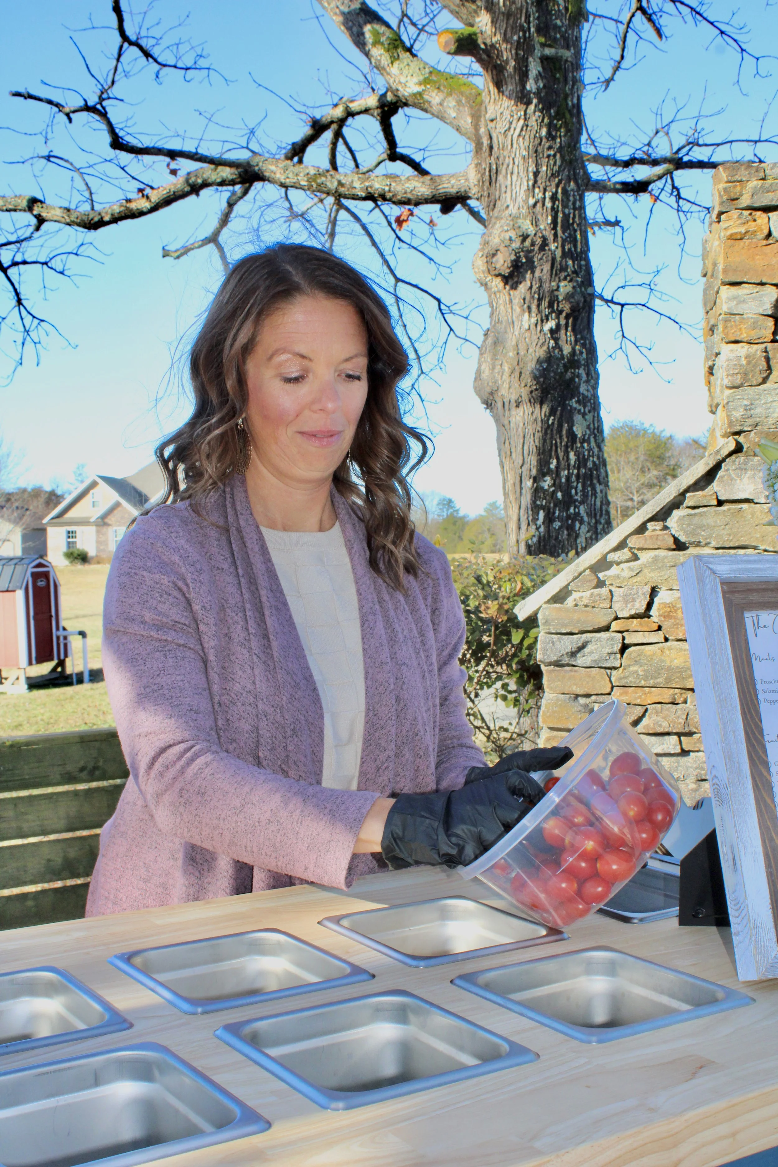 Amber standing by her mobile charcuterie cart wearing black gloves, is holding a clear container of cherry tomatoes for The Charcuterie Chics near Raleigh NC