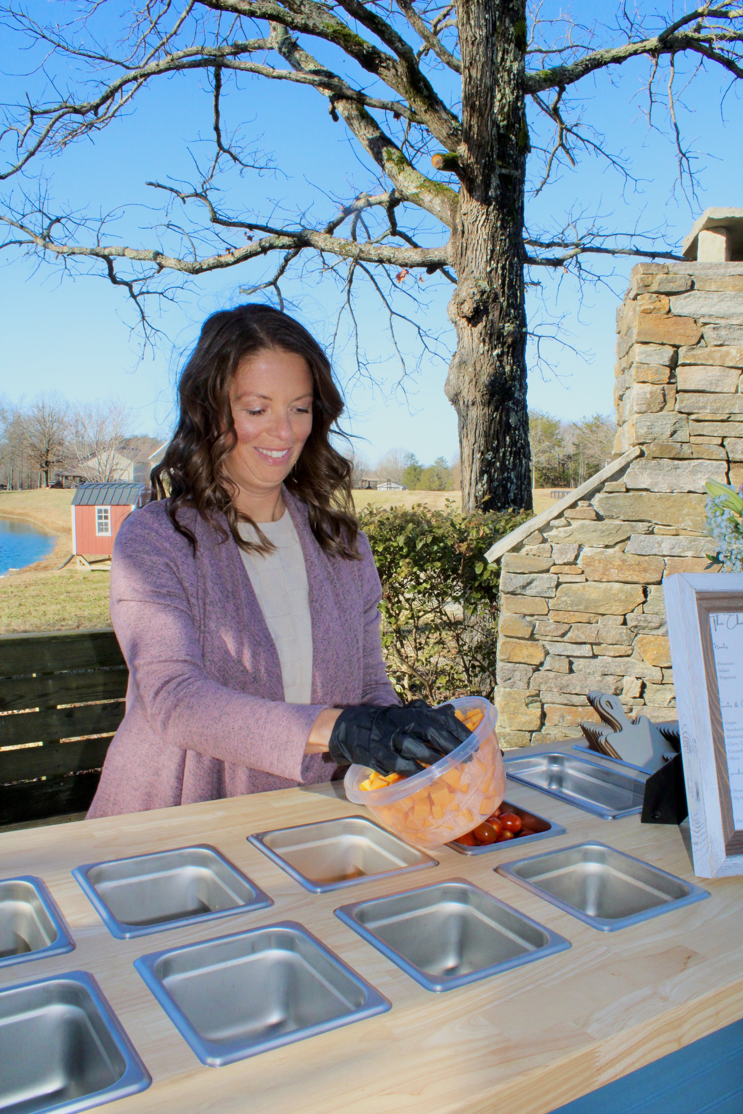 Amber standing by her mobile charcuterie cart wearing black gloves, is holding a clear container of cheeses for The Charcuterie Chics near Raleigh NC