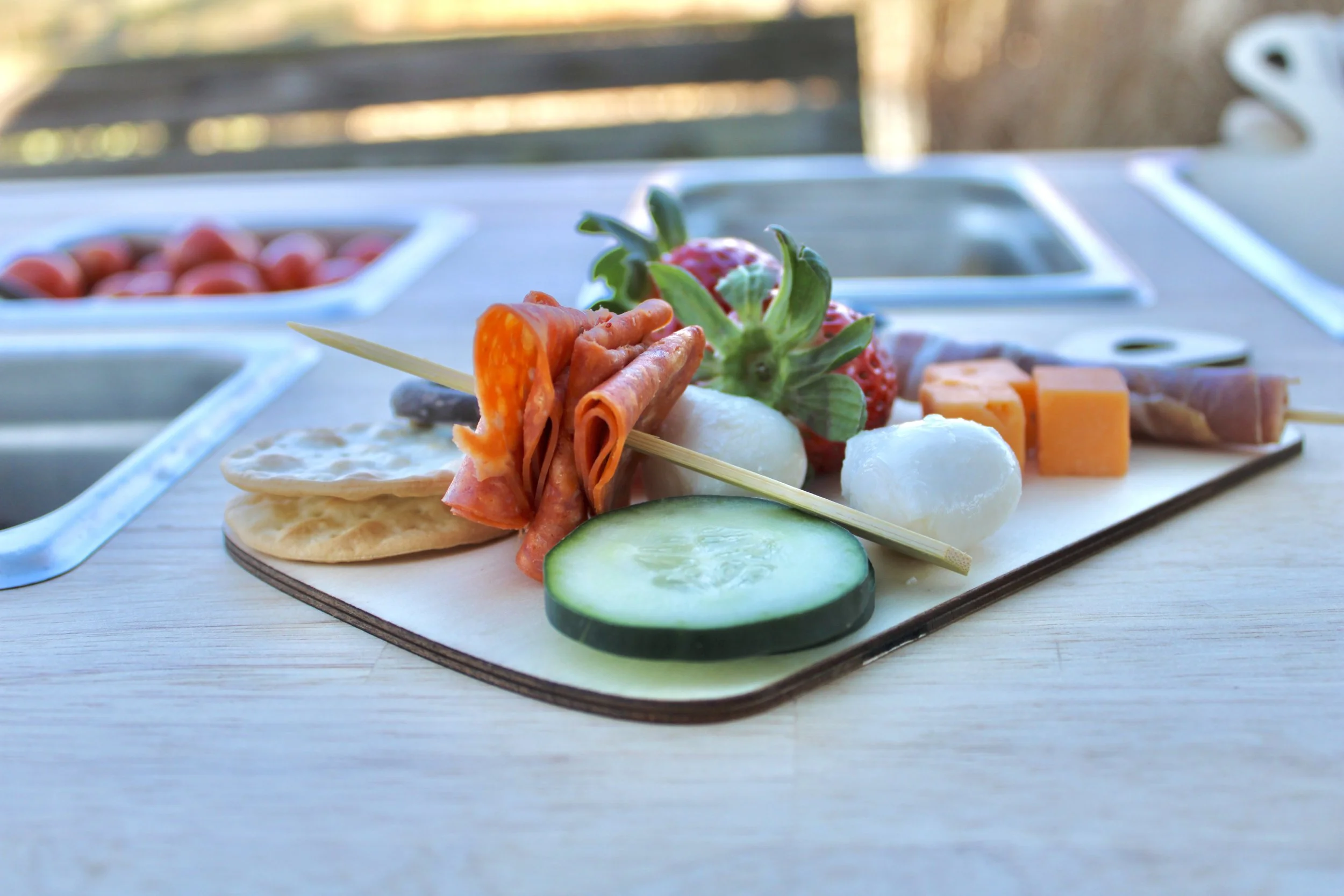 A wooden serving board featuring sliced cucumber, strawberries, mozzarella balls, cheese cubes, salami, and prosciutto, with cherry tomatoes in a bowl in the background for The Charcuterie Chics near Raleigh NC