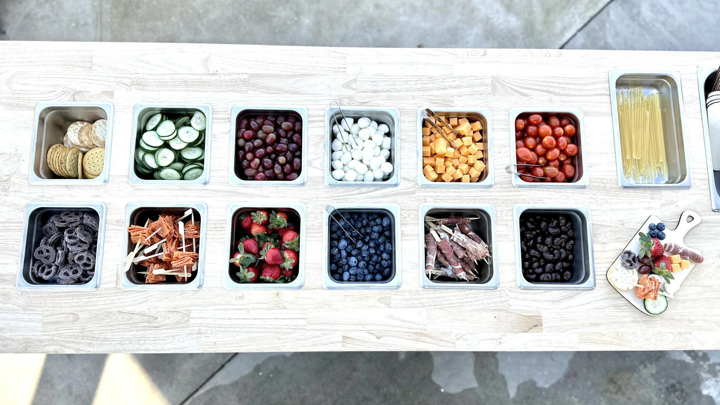 Mobile charcuterie cart with 14 containers of various charcuterie including crackers, cucumber slices, grapes, mozzarella balls, cubed cheese, cherry tomatoes, spaghetti, black licorice, and berries, with individual charcuterie boards near Charlotte 