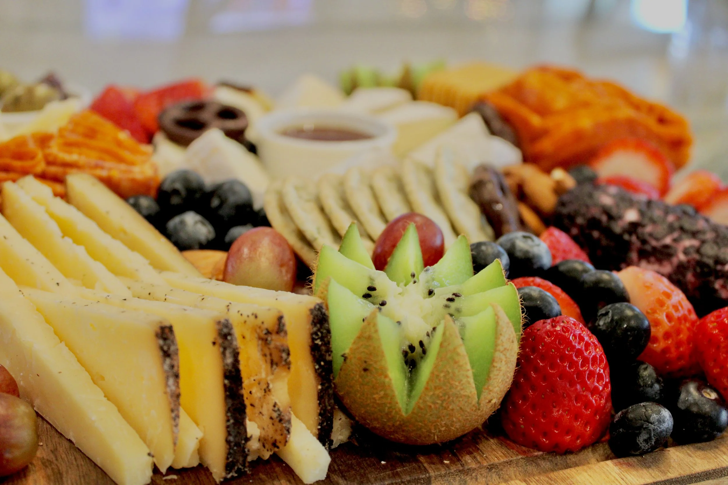 Assorted sliced cheese, grapes, strawberries, blueberries, and sliced kiwi on a wooden board, with some crackers and a small cup of dip in the background from The Charcuterie Chics near Charlotte NC
