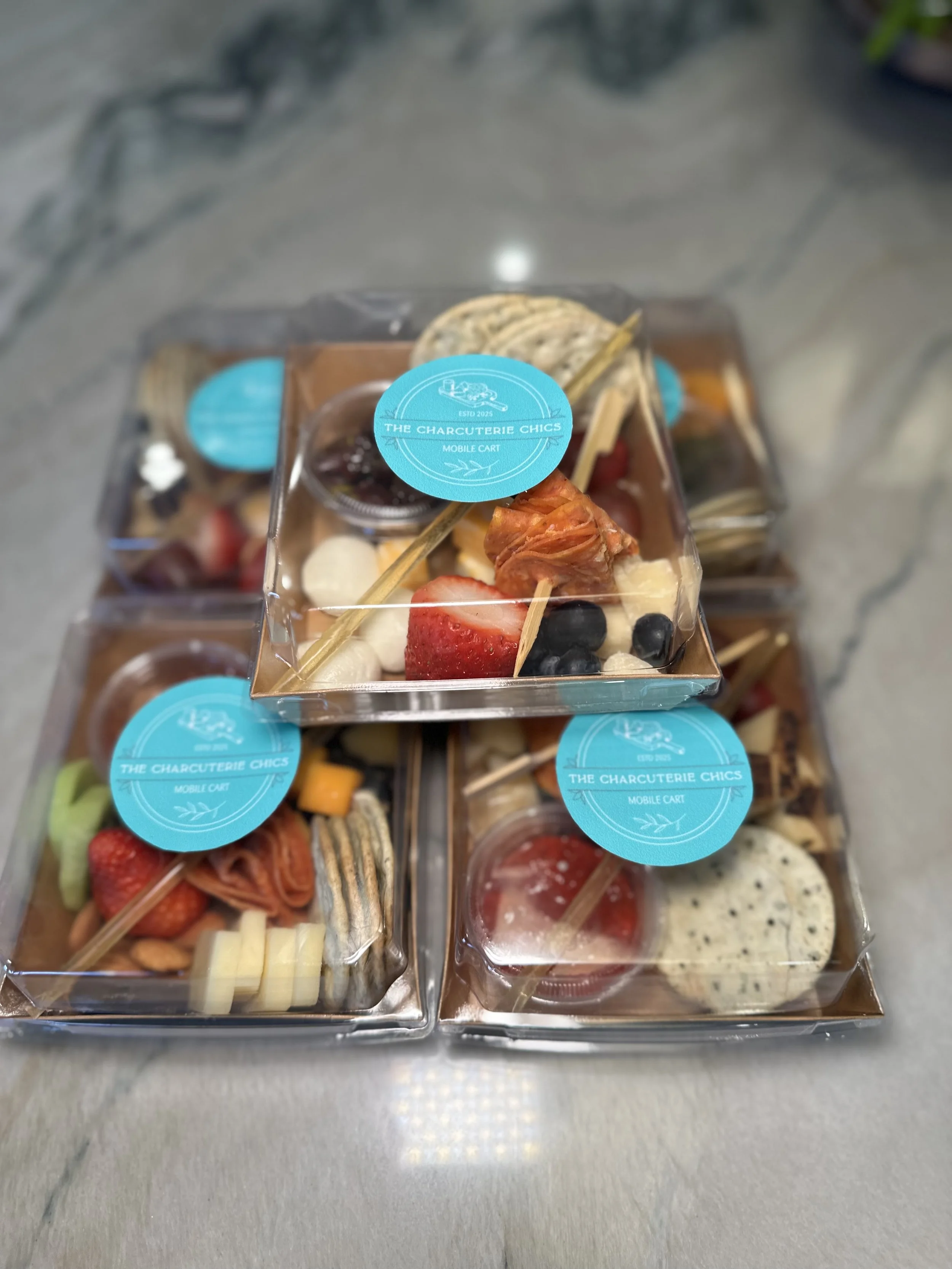 Three to-go charcuterie boxes of assorted charcuterie and fruit snacks with blue labels reading 'The Charcuterie Chics' on a marble countertop from The Charcuterie Chics near Winston Salem NC