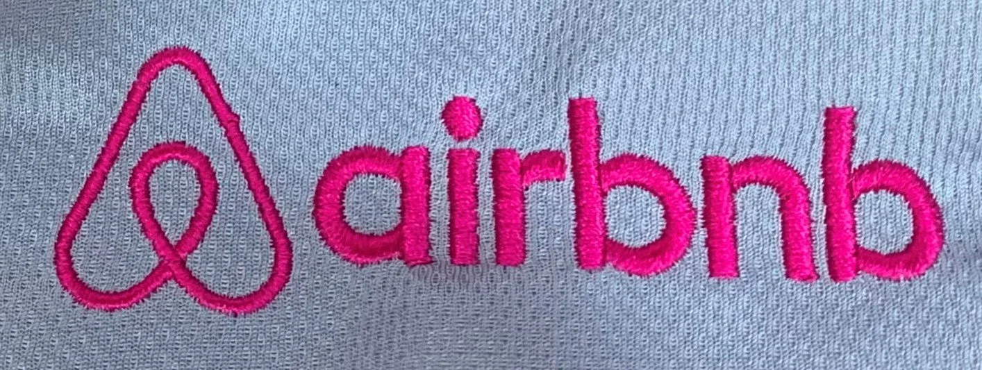 Close-up of pink embroidery on gray fabric showing the Airbnb logo and the word "airbnb".