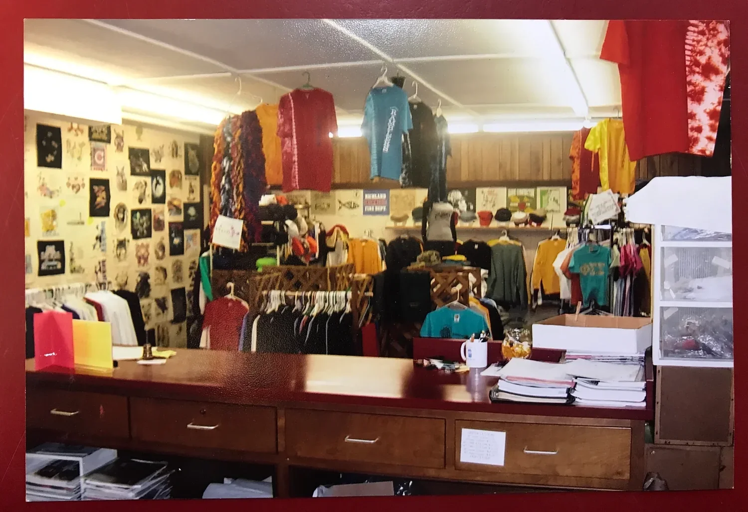 A small retail store with clothing and accessories on racks, walls decorated with art, and a wooden counter with stacks of paper and a mug.