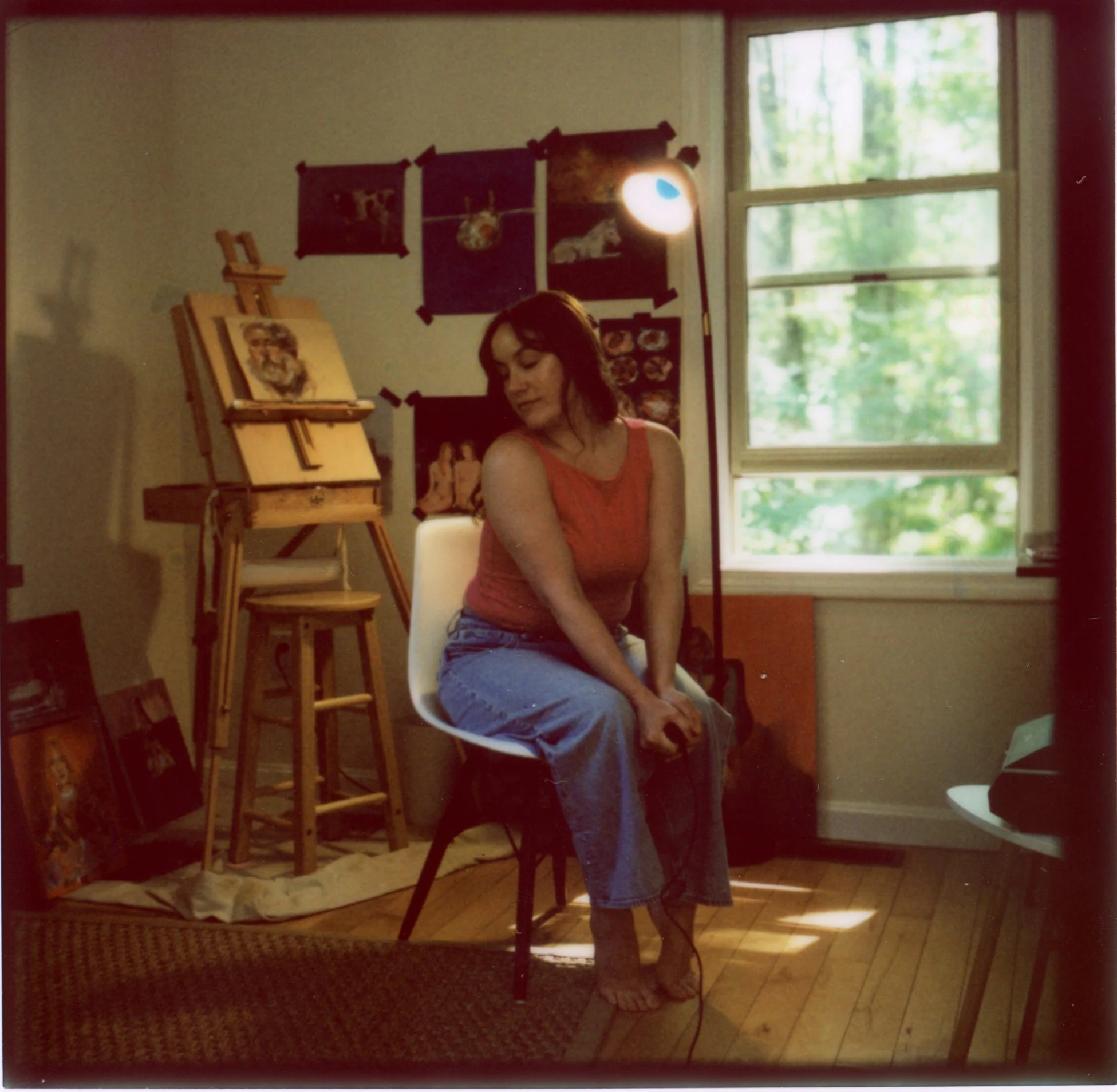 A woman with dark hair sitting by a window in a room decorated with art and photographs. There is an easel with a painting, and a red and beige patterned rug on the wooden floor.