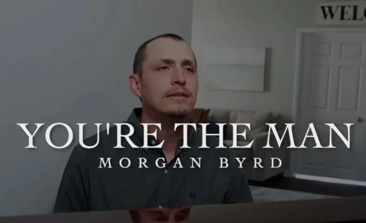 A man with short dark hair, wearing a black shirt, sitting in a room with gray walls. There is a sofa and a door in the background. Overlaid text reads: "YOU'RE THE MAN" and "MORGAN BYRD." The top right corner has part of a sign that reads: "WELCOME.