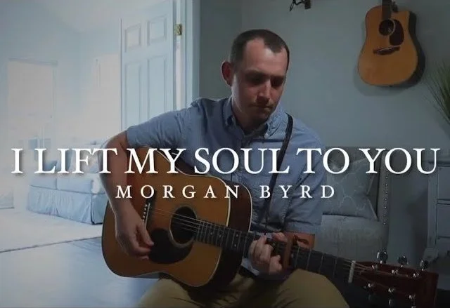 A man playing an acoustic guitar in a room with a blue wall and a guitar hanging on the wall. The text overlay says 'I LIFT MY SOUL TO YOU' by Morgan Byrd.