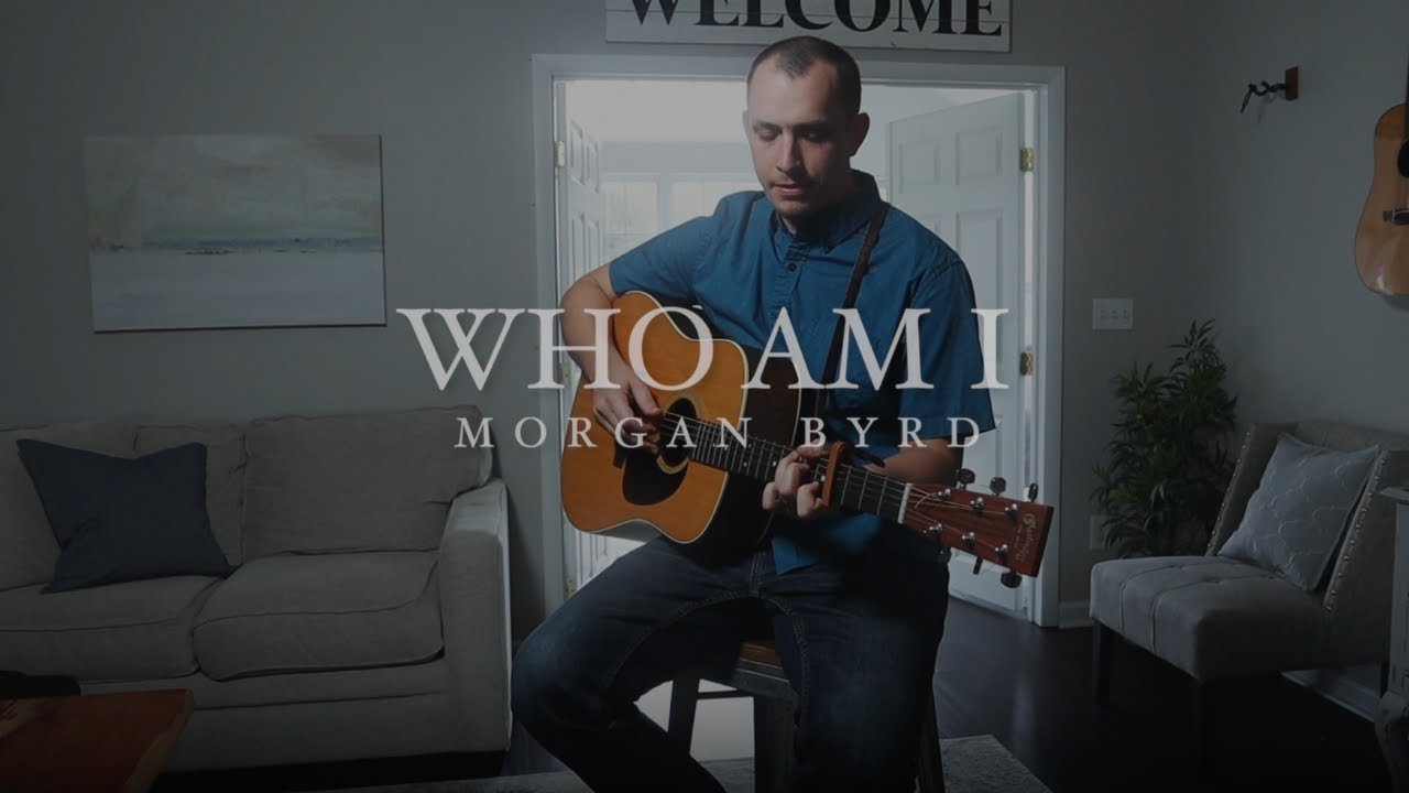 A man playing an acoustic guitar in a living room, with a window in the background and a welcome sign on the wall.