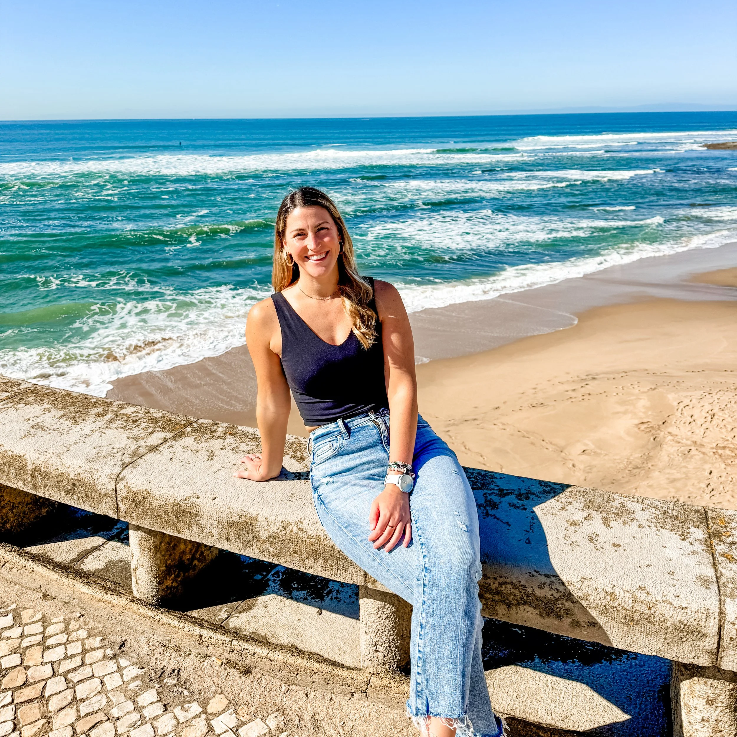 A woman sitting on a stone seawall at the beach, smiling with ocean waves in the background on a sunny day.