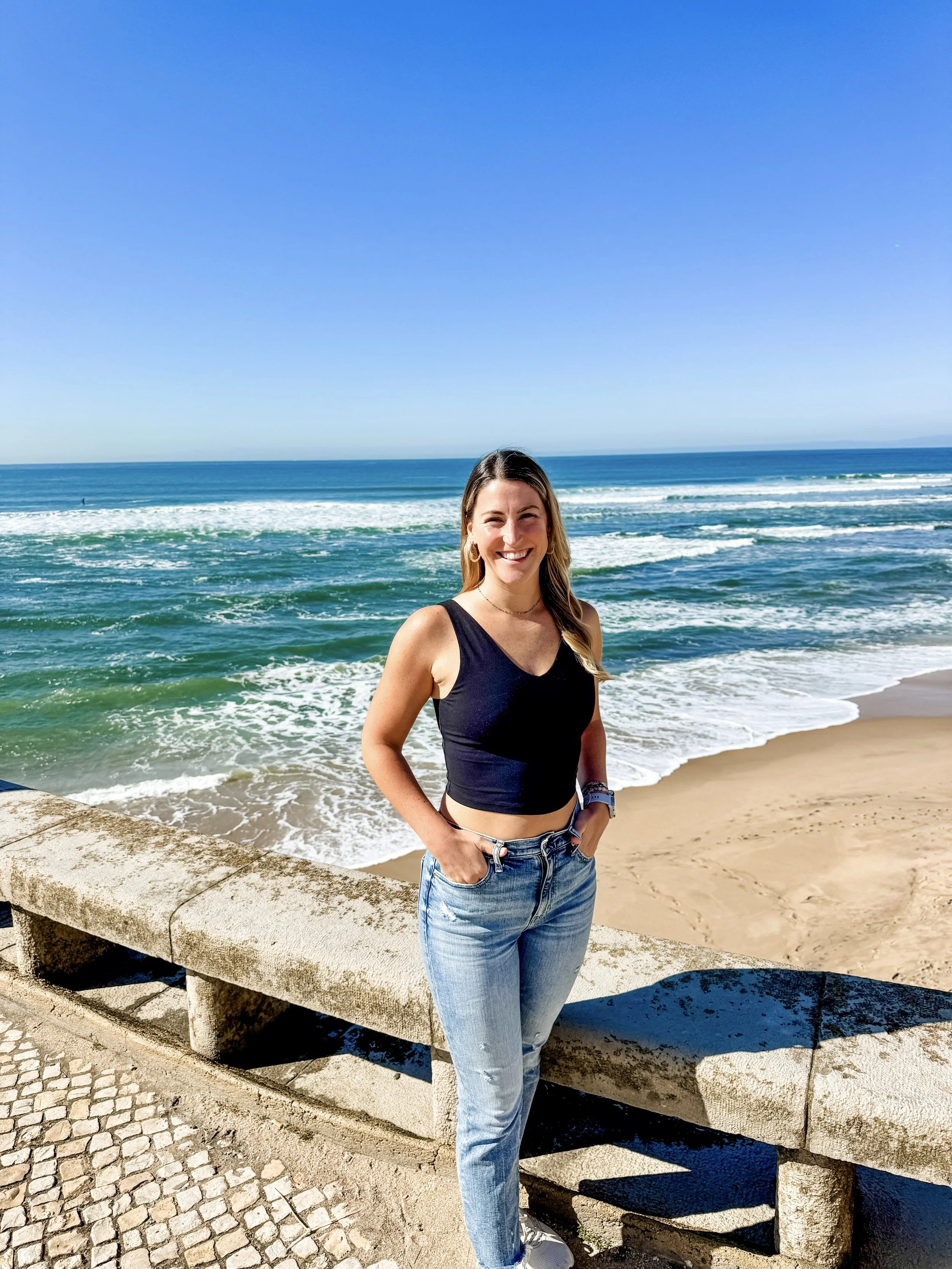 Smiling woman in black tank top and jeans standing on a stone barrier by the beach with ocean waves and blue sky in the background.