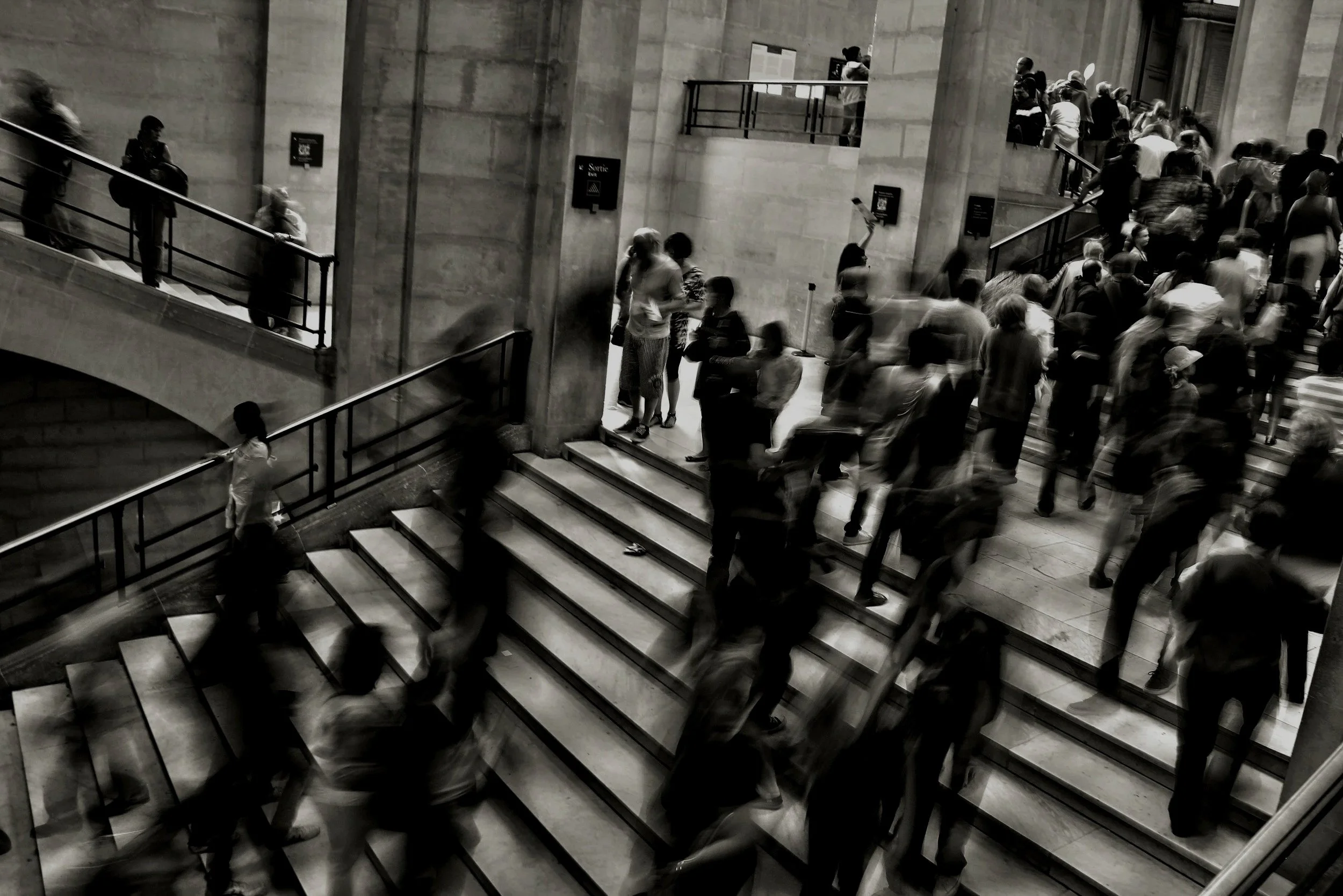 A black and white photo of a crowded staircase in a museum or public building, with people walking up and down the stairs, some standing and conversing, and others looking at exhibits or signs on the walls.